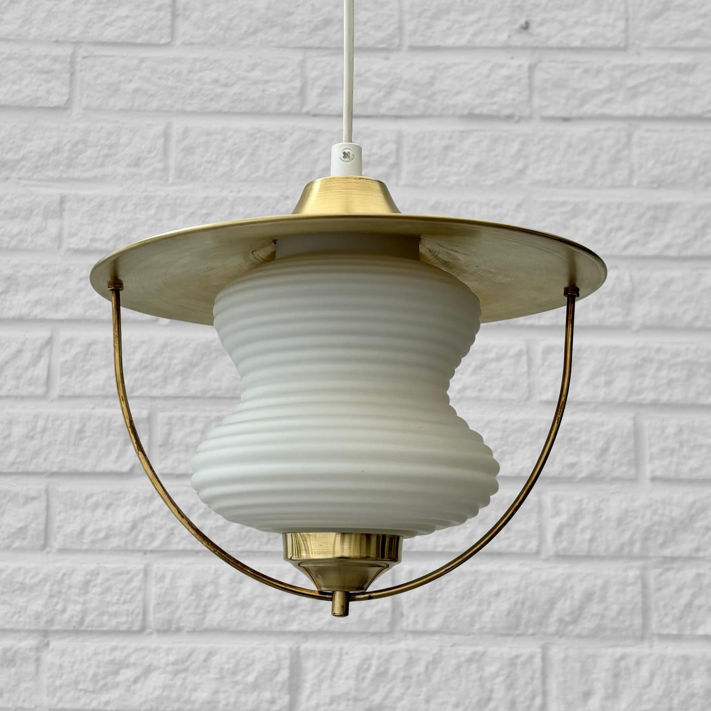 Lampe suspendue en forme de lanterne fabriquée en laiton et dotée d'un abat-jour en verre en forme de cacahuète. L'abat-jour supérieur en forme de disque est maintenu en place par deux vis décoratives en forme de sphère. Elle est suspendue par un