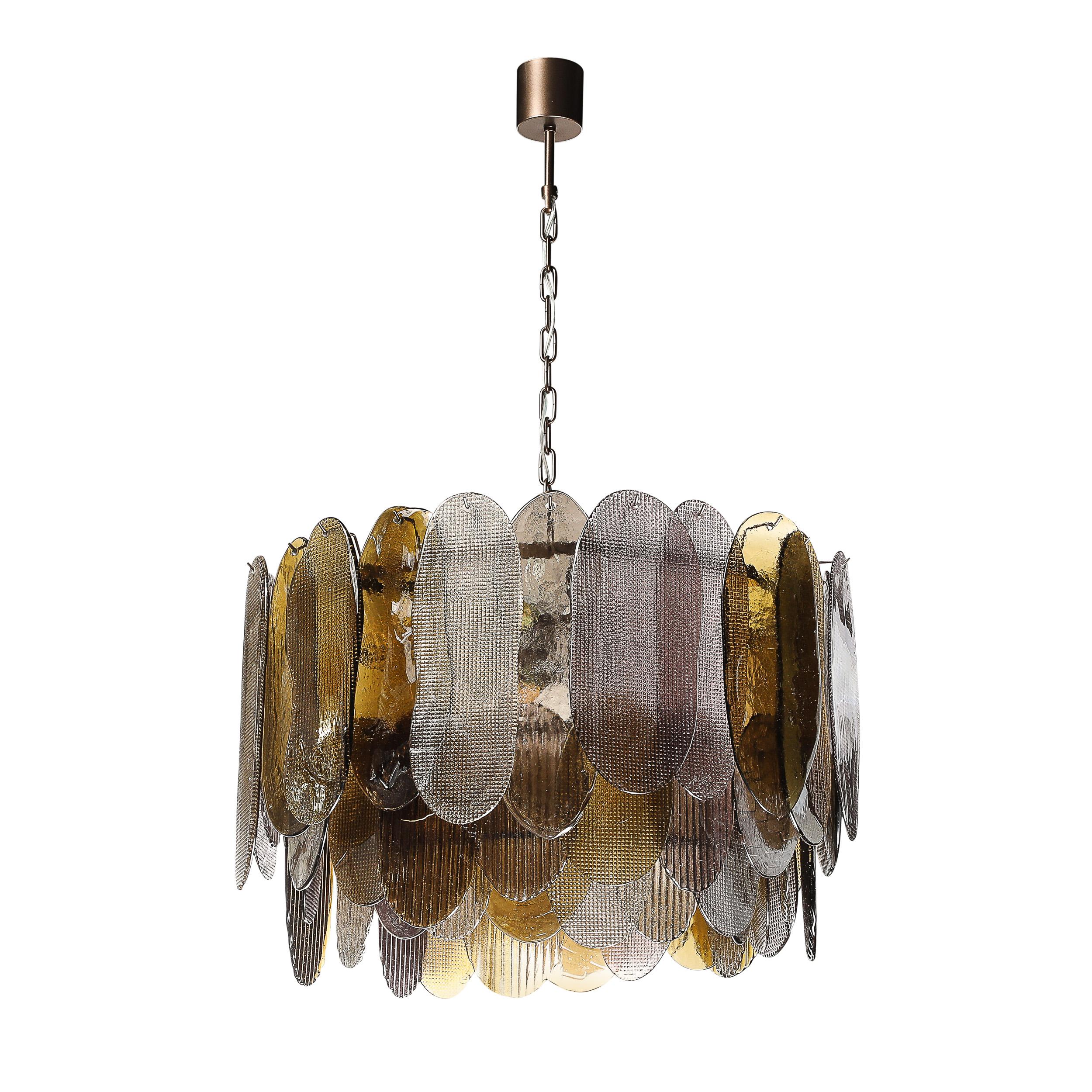 Ce lustre moderniste exquis du XXIe siècle présente trois niveaux de verre de Murano soufflé à la main, mettant en valeur le savoir-faire et l'art intemporels des verriers italiens. Le lustre est orné d'abat-jour ovales cannelés et hachurés dans une