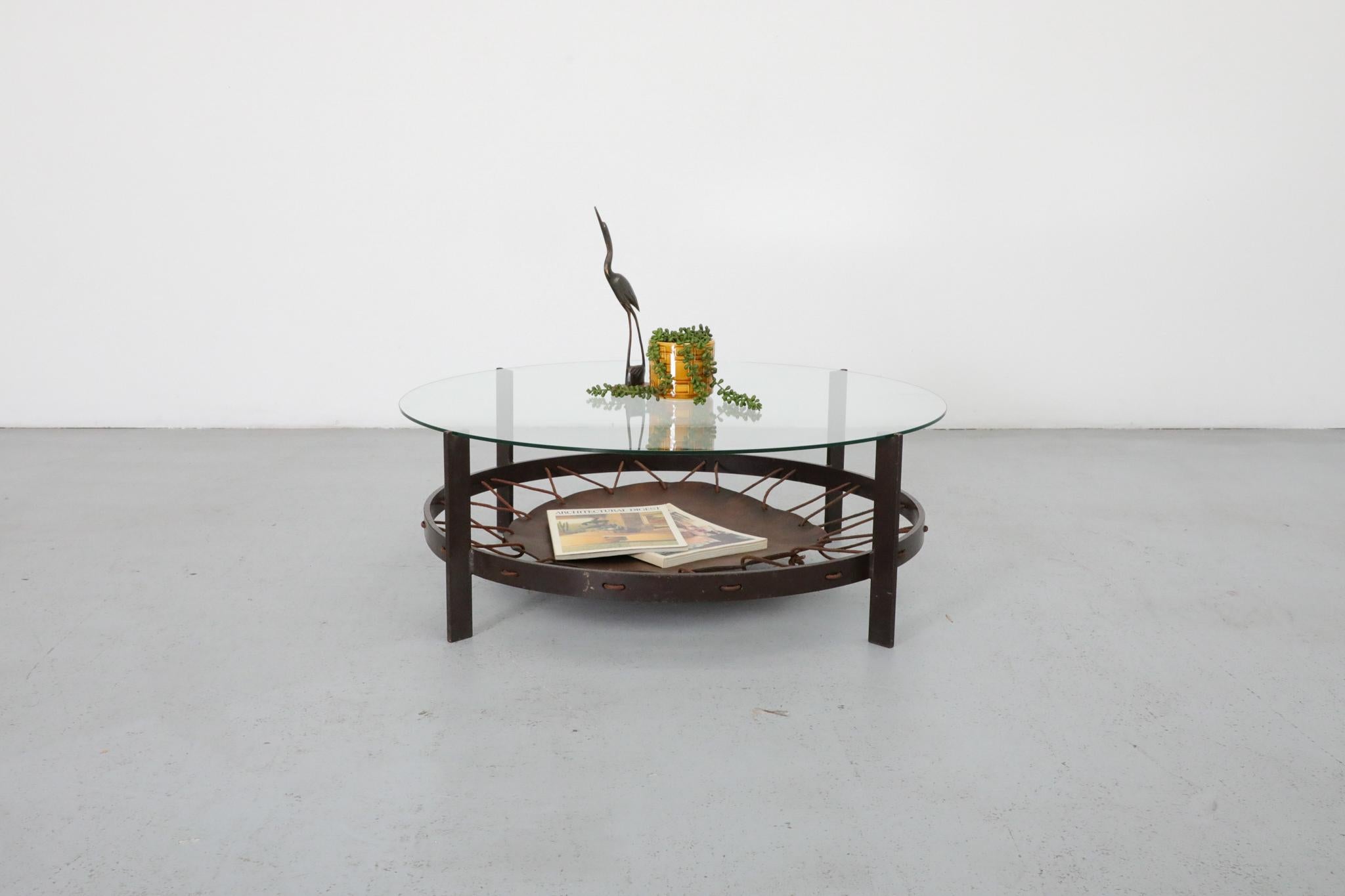 Striking Modernist coffee table featuring a round glass top supported by a flat-bar metal frame finished in a warm brown enamel. The true standout detail lies beneath: a suspended leather magazine sling, richly patinated from age and use, held in