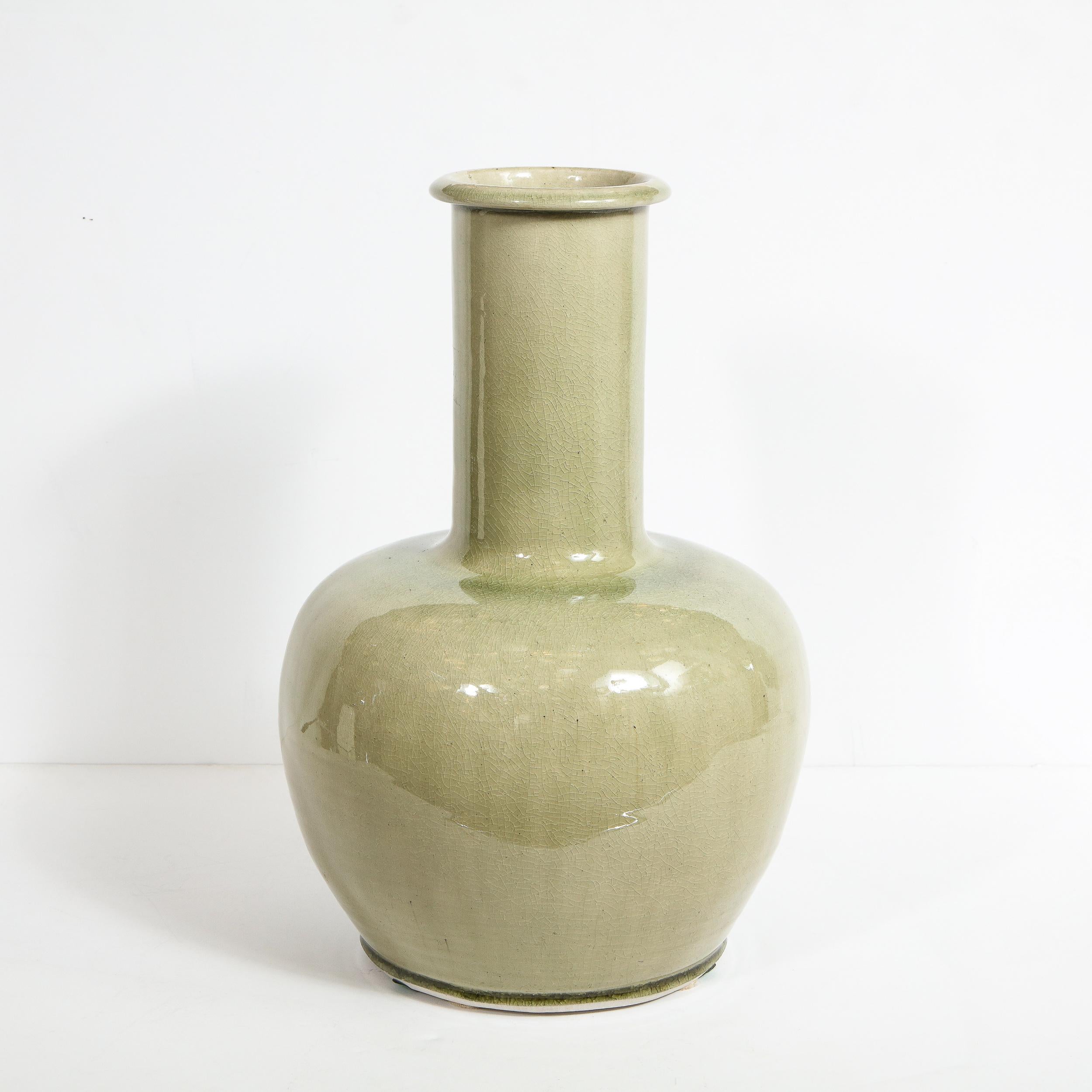 This elegant modernist vase features an organic spherical bottom that tapers to its base, and elongated cylindrical neck, and a raised lip around the circular mouth. The piece offers a lovely craqueleur finish in a sultry sage hue. With its refined