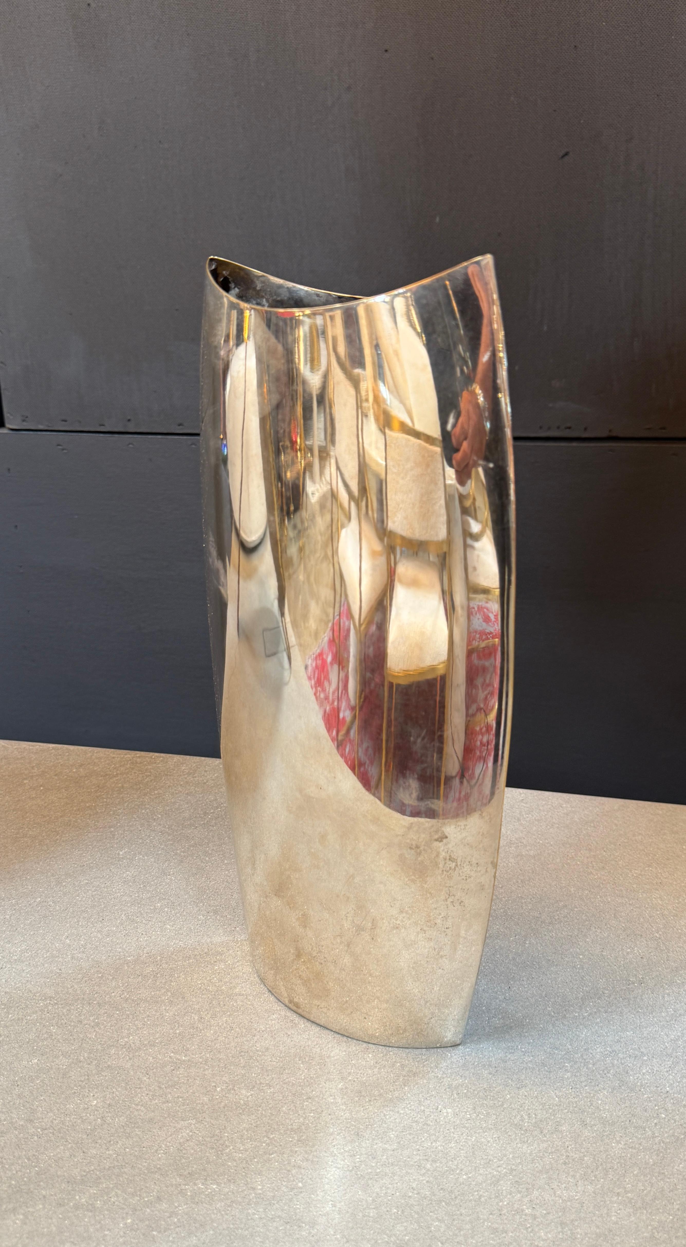 Questo sorprendente vaso modernista presenta un design elegante e asimmetrico con superfici argentate lisce e riflettenti. La sua forma curva e organica cattura e distorce la luce e i colori circostanti, creando un effetto visivo dinamico. Ideale