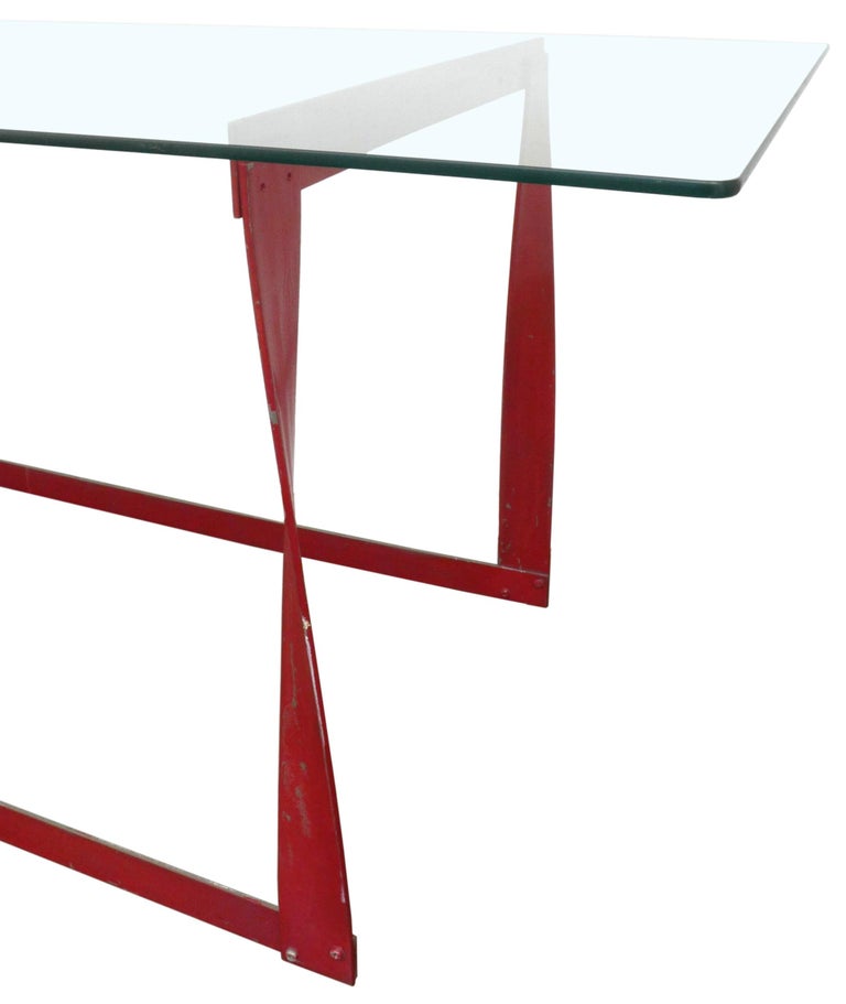 An unusual, modernist, painted steel and glass dining table. A simple rectangular glass top rests upon a graphically striking base of red-painted, flat-stock steel that has been twisted into form. Beautifully constructed and bold in its playful