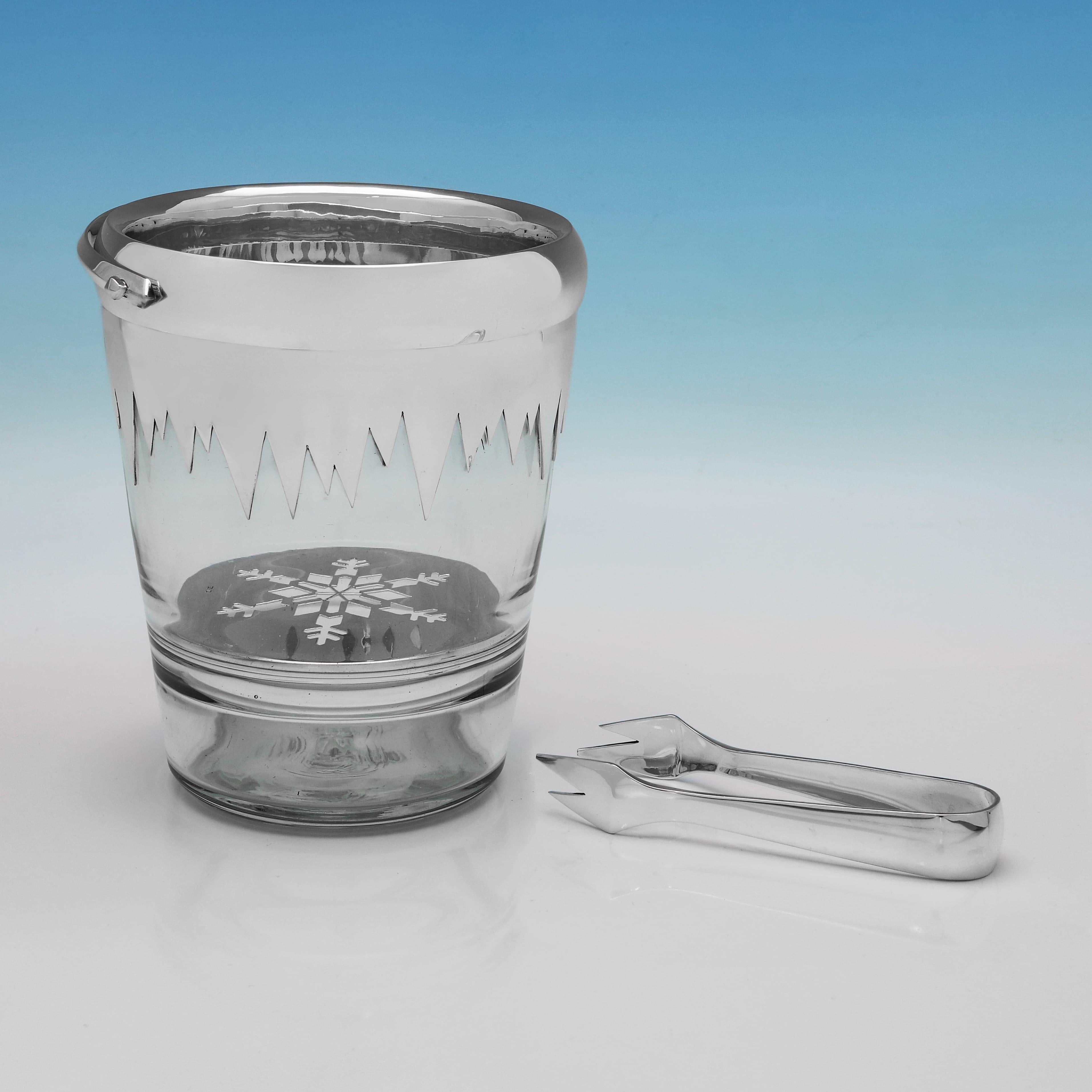 Hallmarked in London in 1988, this sterling silver and glass ice bucket was made by James W. Powell. The cylindrical glass body is topped with a broad silver mount, shaped in a jagged, icicle-like form. A swing handle is fitted to either side. At