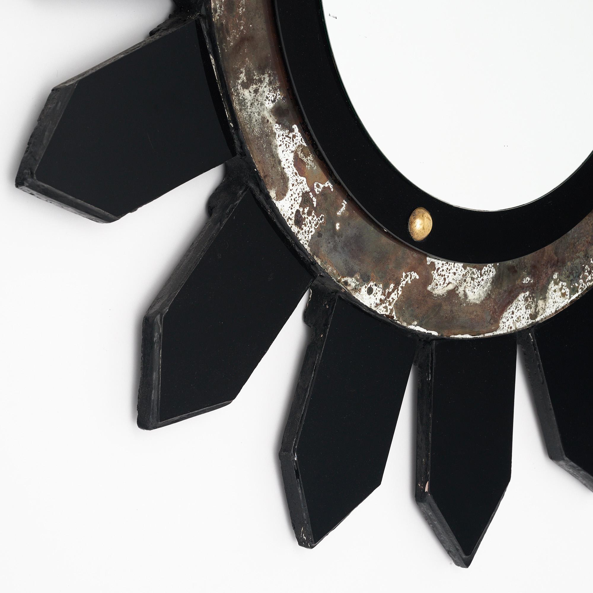 Modernist sunburst mirror made with a wooden structure and a central circular mirror. The rays are all covered with black glass. The central mirror is framed with antiqued mirror and black glass with gold leafed details. We love the stylized look of