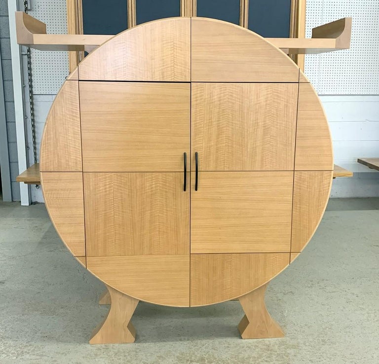 Custom sycamore cabinet of circular form on four shaped legs with double doors which open out as well as slide back into the cabinet. On top of the cabinet is a mounted horizontal shelf that is removable. Finished on the back for placement in the