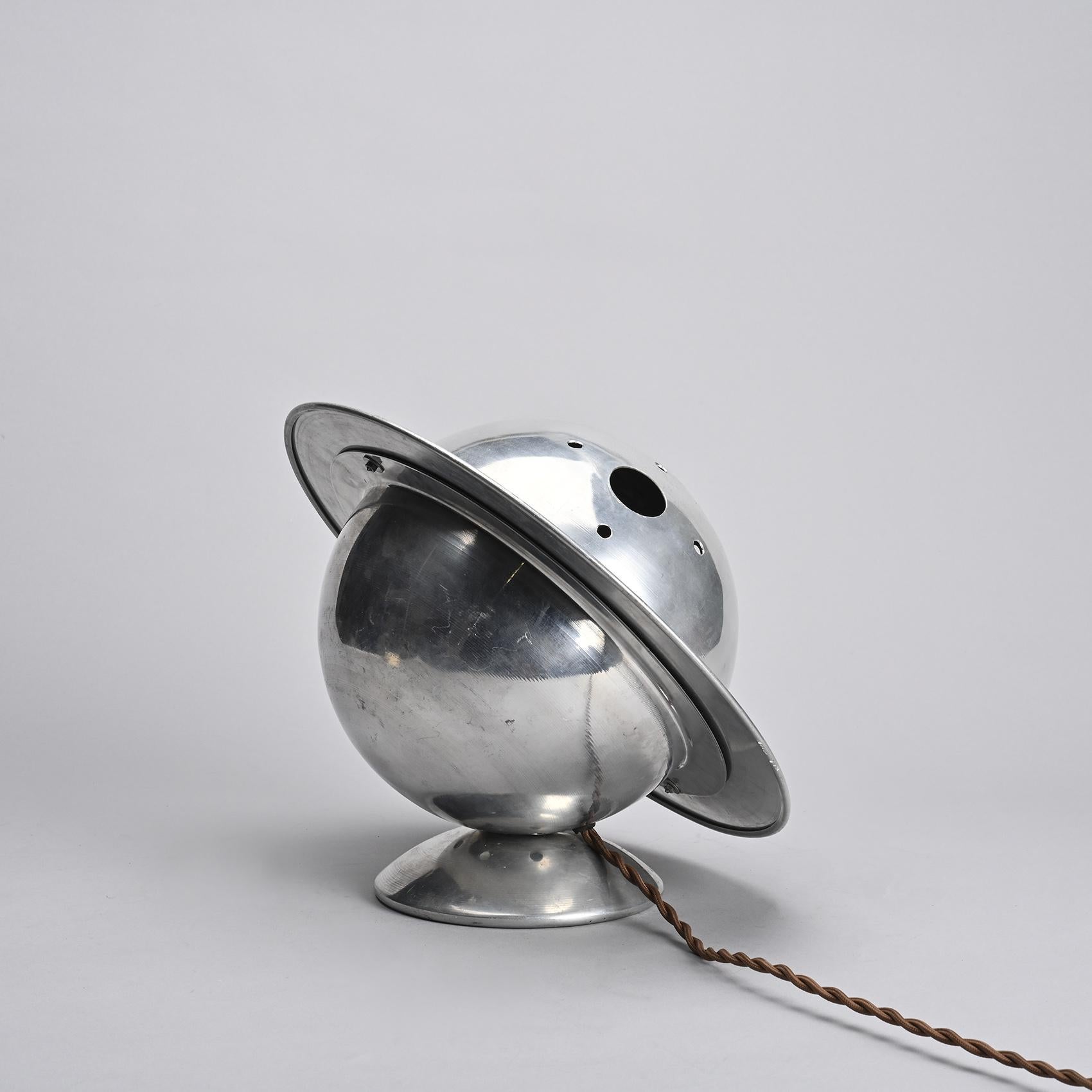 
Sculptural table lamp in nickel-plated aluminum, featuring a wide circular shade tilted around a hemispherical reflector. The overall form suggests a planetary silhouette, reminiscent of Saturn, a motif often explored in modernist lighting design