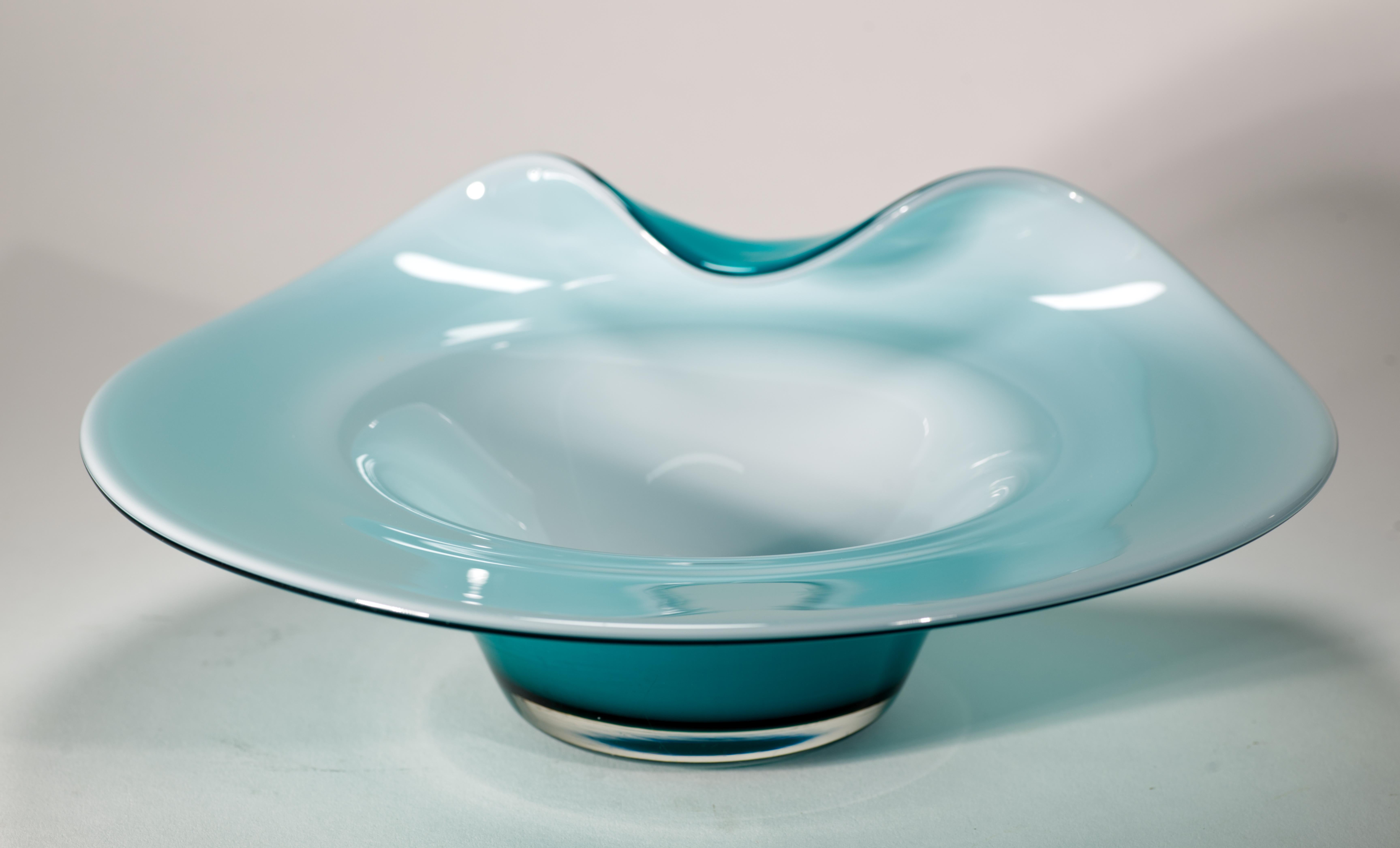  Biomorphic bowl with asymmetrically folded side features a combination of teal blue and white colors. The white glass is encased in teal blue glass in sommerso technique, with some color showing through the thin, semi-translucent layer of white