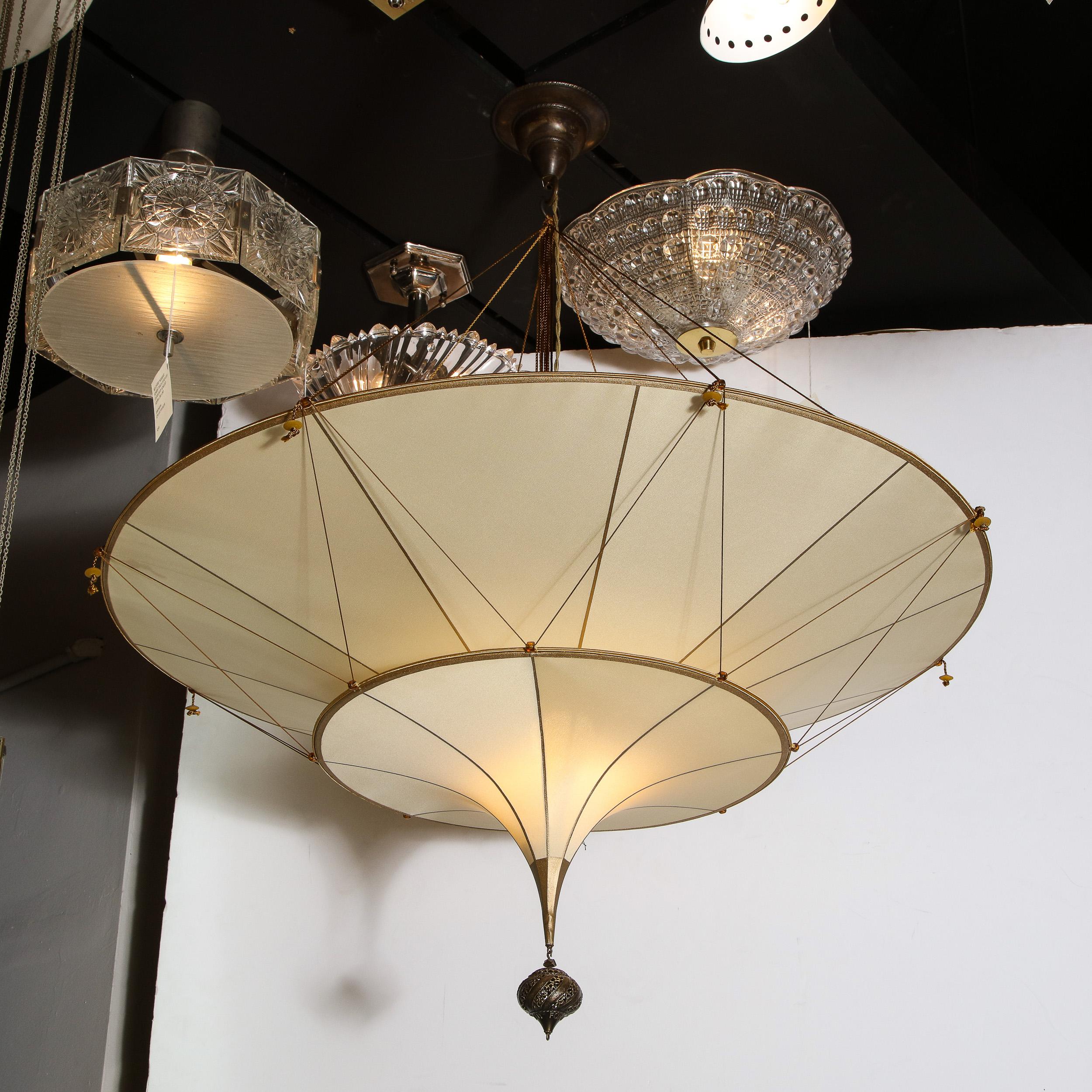 This elegant Mid-Century Modern Scheherazade chandelier was realized by Mariano Fortuny in Italy. It offers two convex tiers of beige silk with an oil rubbed bronze tapered cylindrical finial that connects to a curved striated and textured tear drop