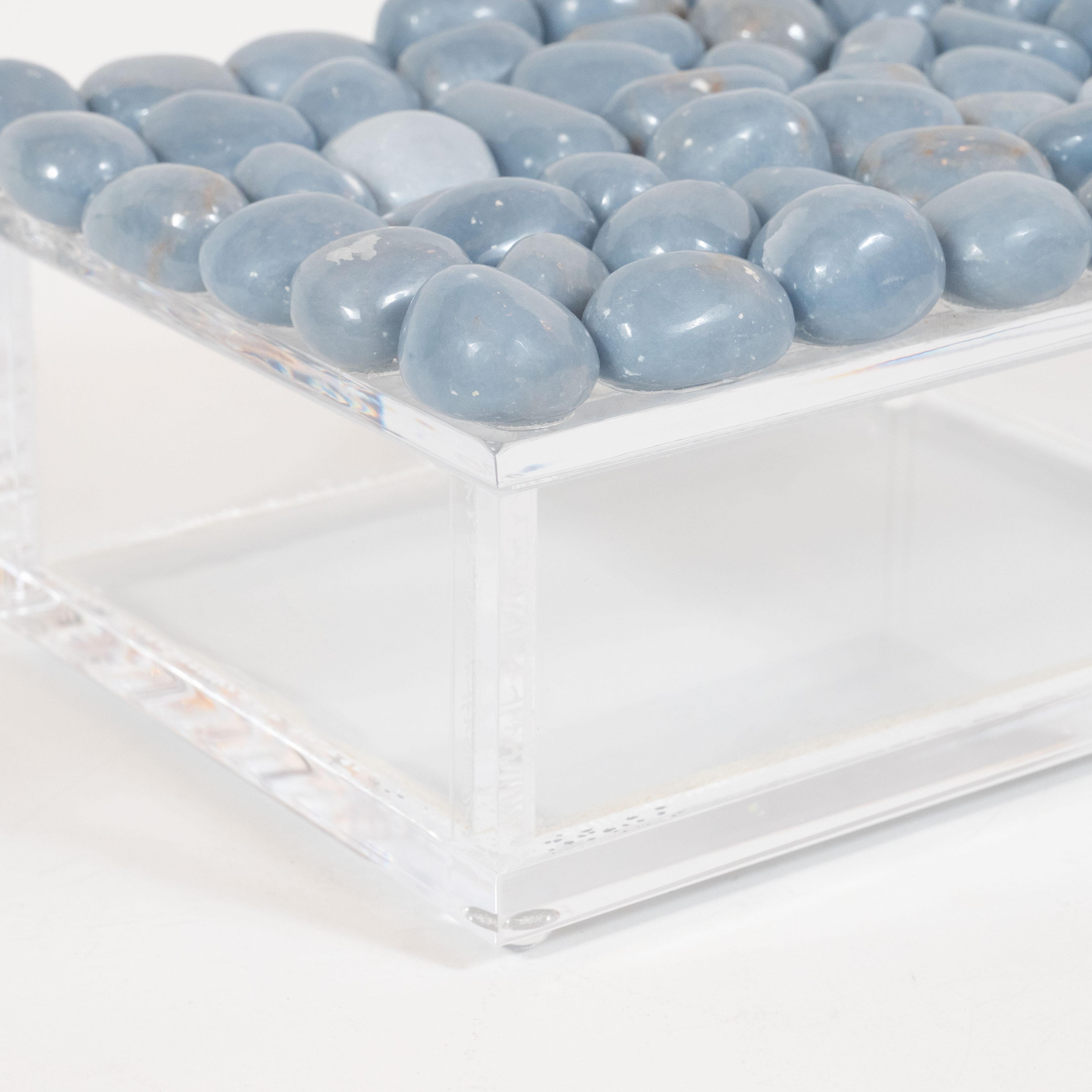 This elegant and unusual features a volumetric rectangular body sitting on orbital feet all realized in clear Lucite. The Lucite top of the box is decorated with a wealth of polished organically formed stones in a muted grayish blue tone, offering a