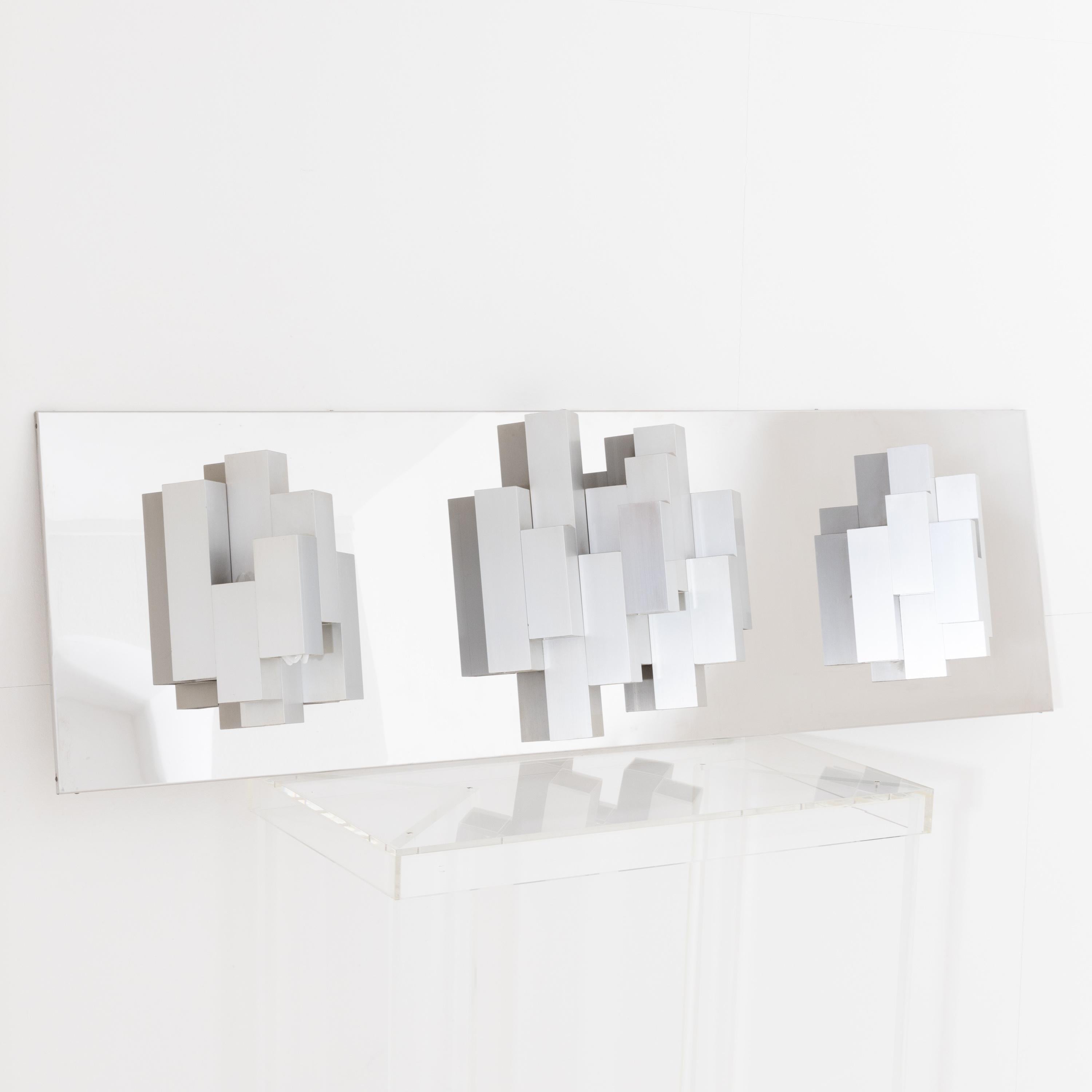 Modernist wall lamp made of nickel-plated metal with clustered cuboids arranged in three groups on a long rectangular base plate. The lamp holders are arranged alternately upwards and downwards, thus creating an interesting play of light.
For the