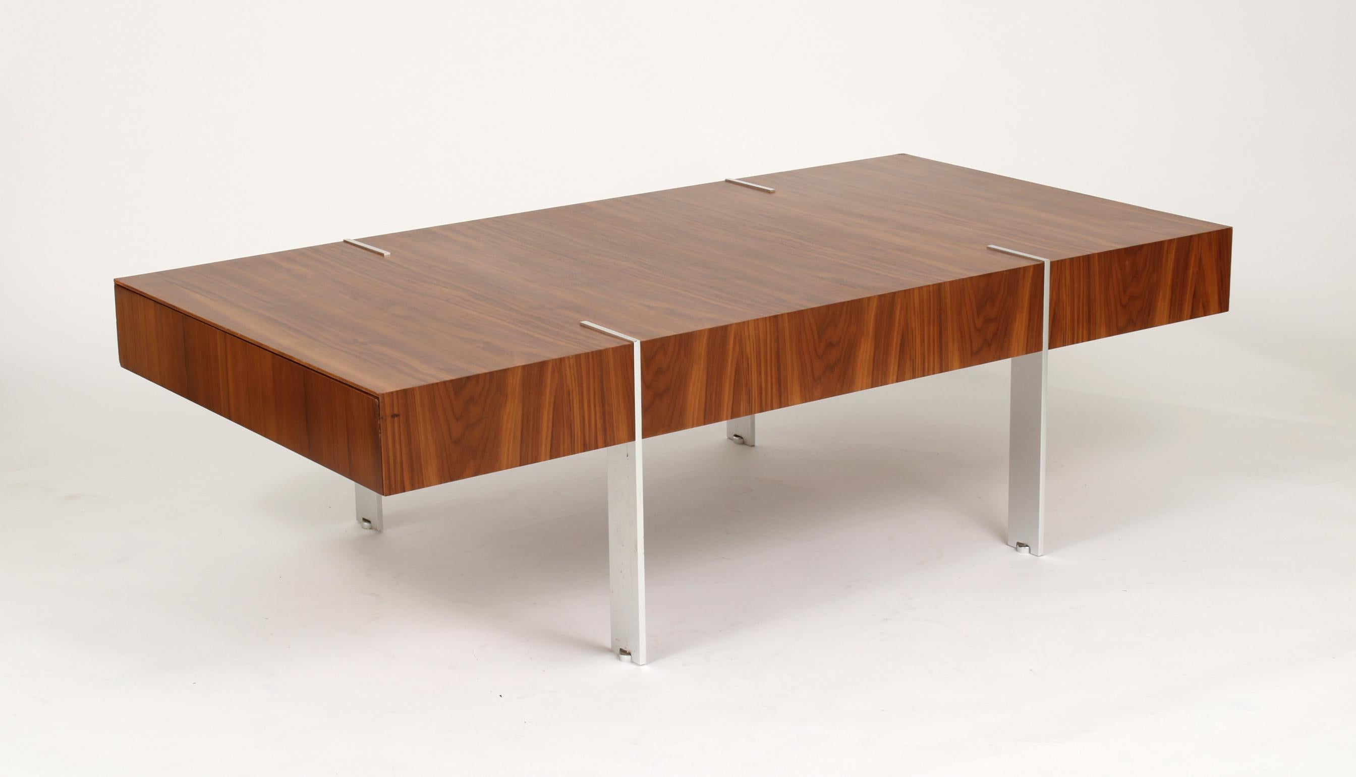 Modernist walnut rectangular coffee table, the top and sides with bookmatched veneer; the ends with concealed drawers; the plated legs continue through the top and terminate with adjustable disc feet.

There are no marks or labels. The carcass is