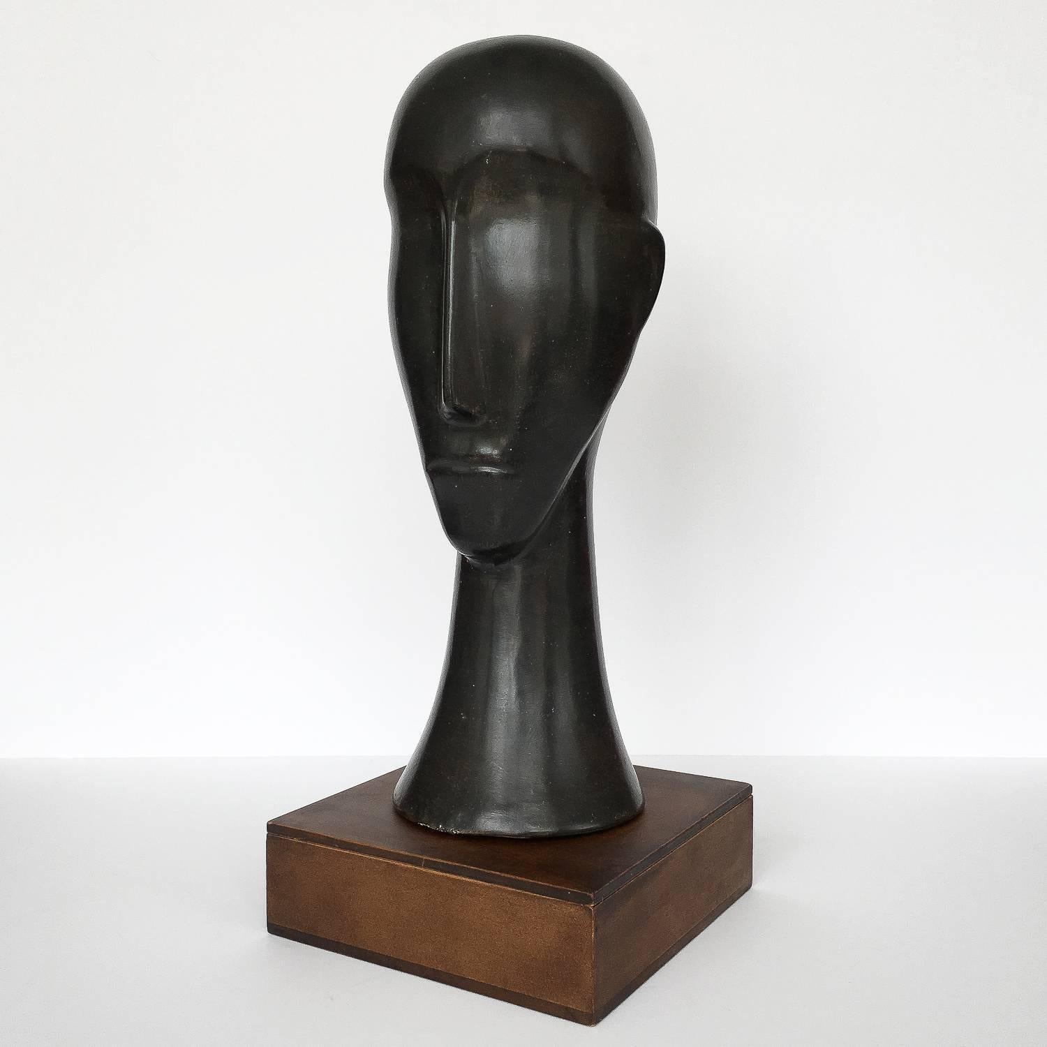 A Modernist Amedeo Modigliani style ceramic sculpture of an elongated male head / bust, circa 1960s. Monolithic in form the ceramic head is glazed in black and is atop a masonite box platform. The sculpture is not affixed to the base. Base measures