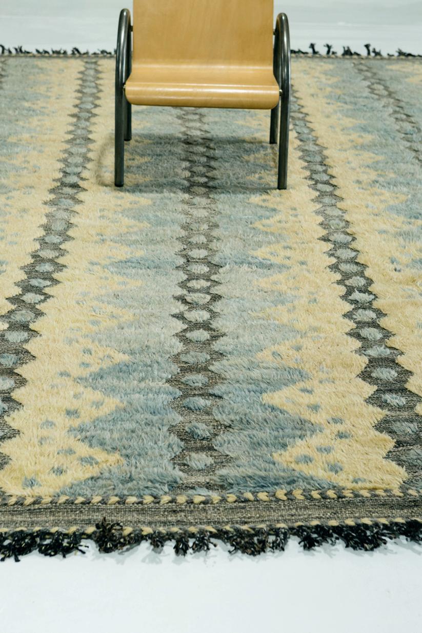 The 'Modlina' rug is a handwoven wool piece inspired by vintage Scandinavian design elements and recreated for the modern design world. The rug's shag balance and harmony, handwoven with a taupish-gray flat-weave and piles of blue and pale yellow.