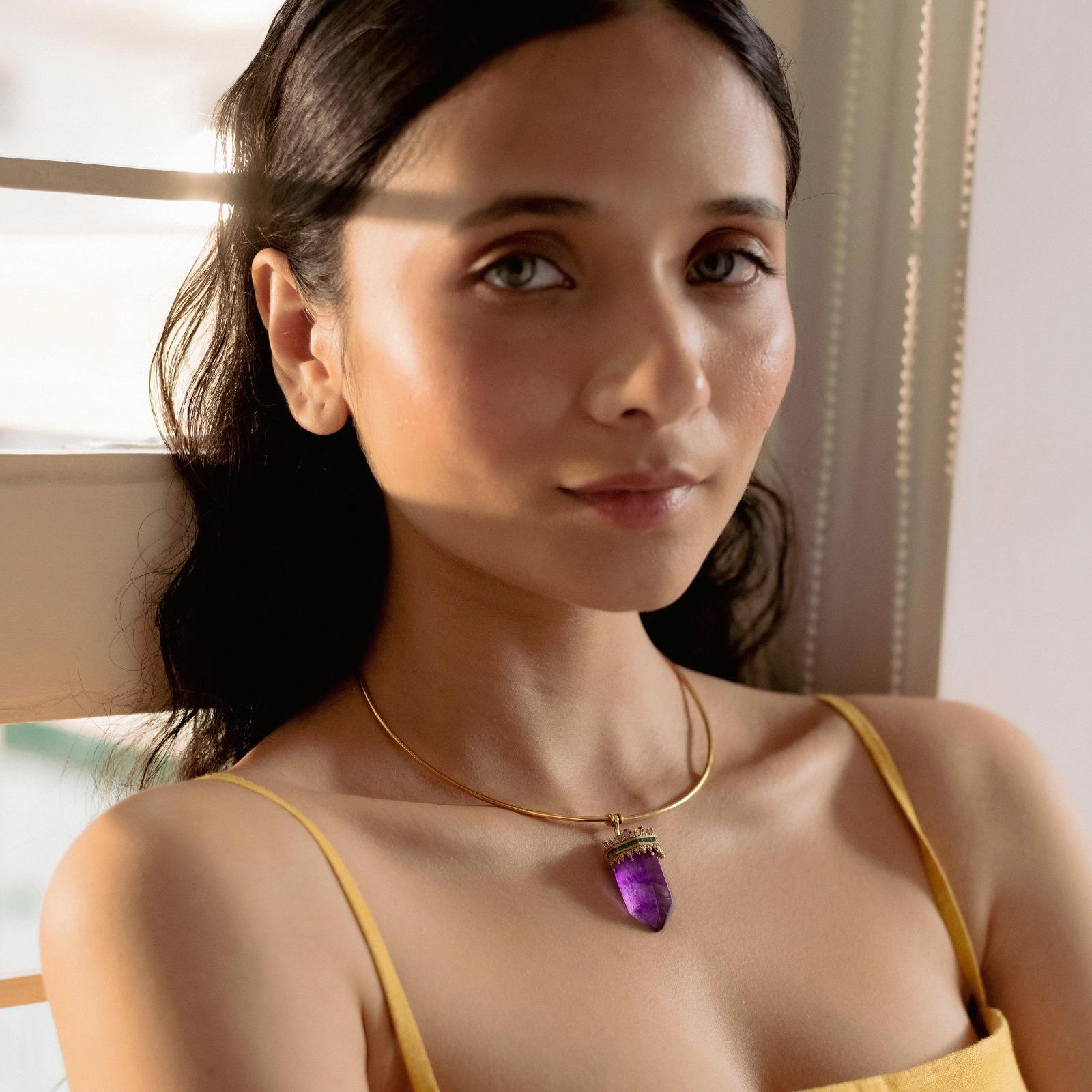 The Promise Torque Necklace strikes a chord with the whimsical. The pendant is a purple amethyst, cut to resemble a crystal and dressed in a tiara of diamonds and emeralds. It is suspended from a thin omega chain/omega hoop, crafted in yellow gold.