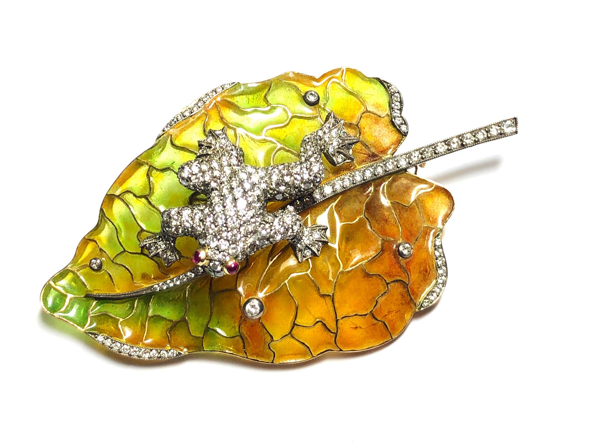 A frog on a leaf brooch designed by MOIRA, with a pavé diamond set frog, with cabochon cut ruby eyes, sitting on a green and ochre plique à jour enamel leaf, set with round brilliant-cut diamonds, mounted in gold, with silver settings. Signed Moira,
