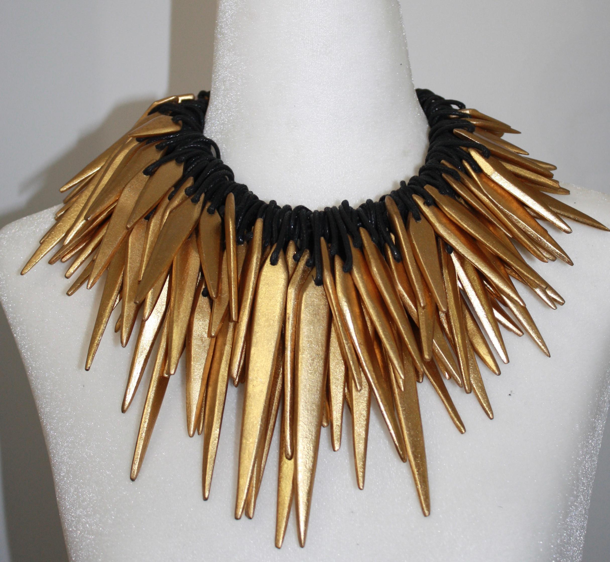 A multitude of spikes in acacia wood covered in gold foil with leather loop are threaded through rows of leather.
The pieces  can be moved around  the leather cord for different looks. The clasp is made of eight strands of leather that can tie