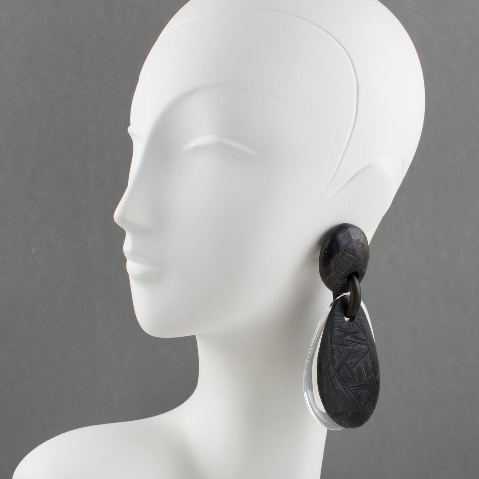 These spectacular oversized clip-on earrings by Gerda Lyngaard for Monies feature a dangling pebble double teardrop shape built with transparent Acrylic or Lucite and Ebony wood with tribal carved texture. The pieces are marked 