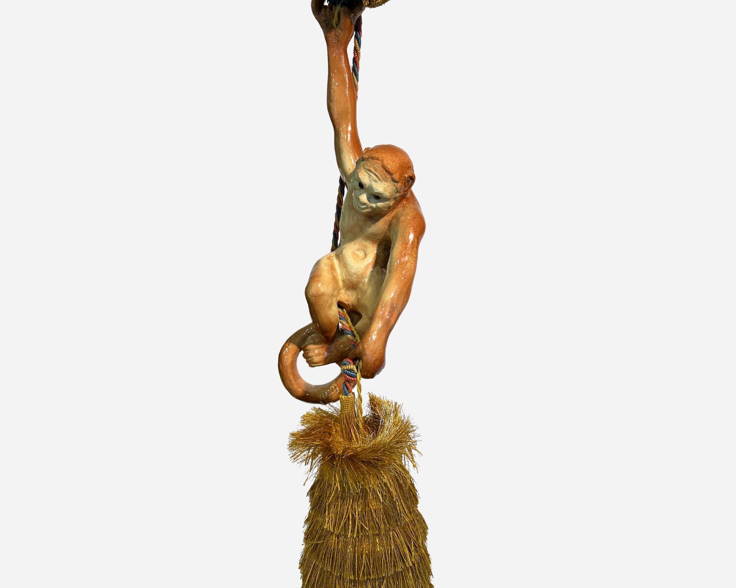 Rare glazed terracotta pendant featuring a small monkey with glass sulfide eyes, holding a ring with a sliding cord in one hand and a (recent) fringed shade containing a light bulb in the other.

This little monkey is probably a production of the