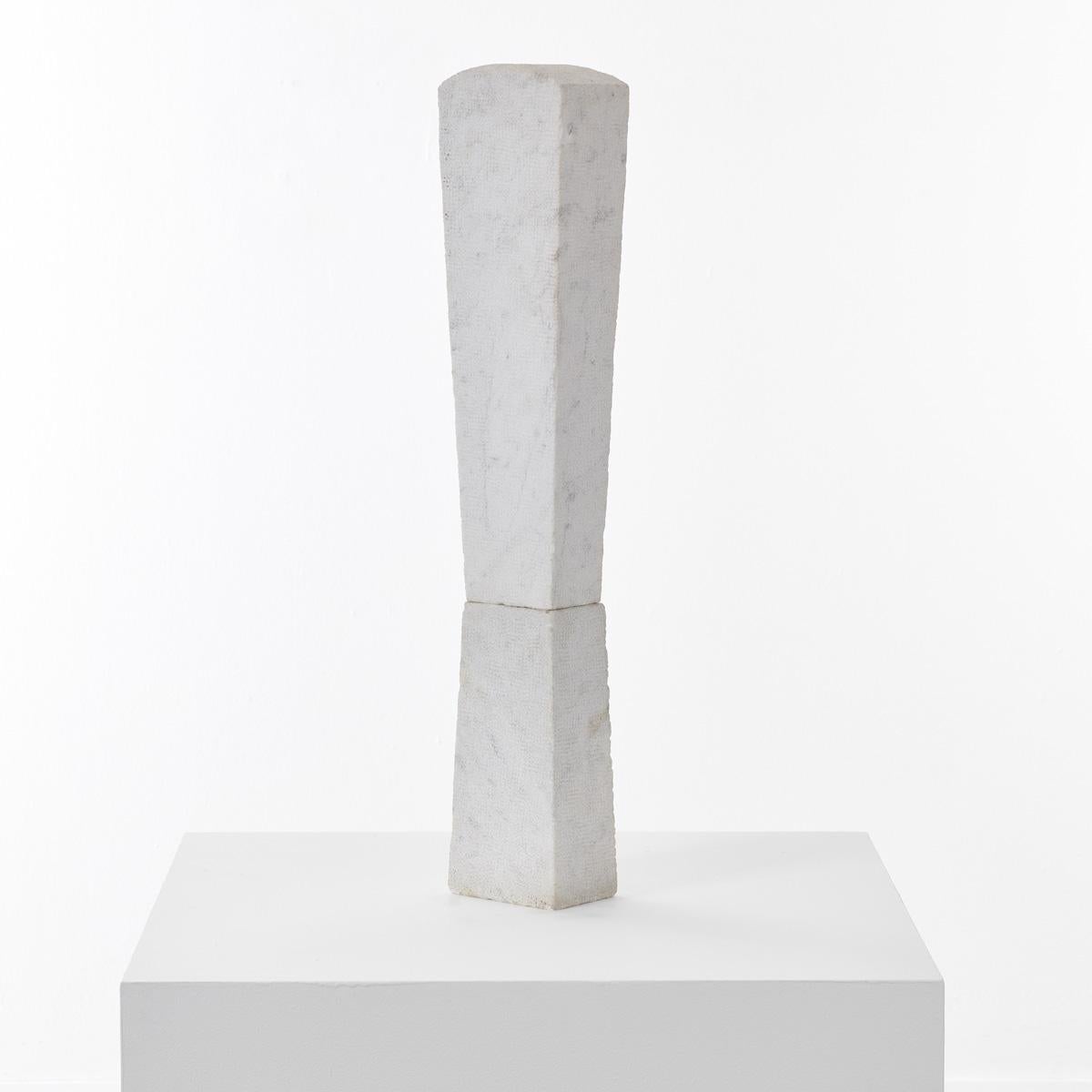 A towering sculpture hand carved from marble in two parts. The top part has had its surfaces finely chiselled to create a fine surface texture. The bottom element is chiselled more roughly in a linear pattern. A metal rod running through the centre