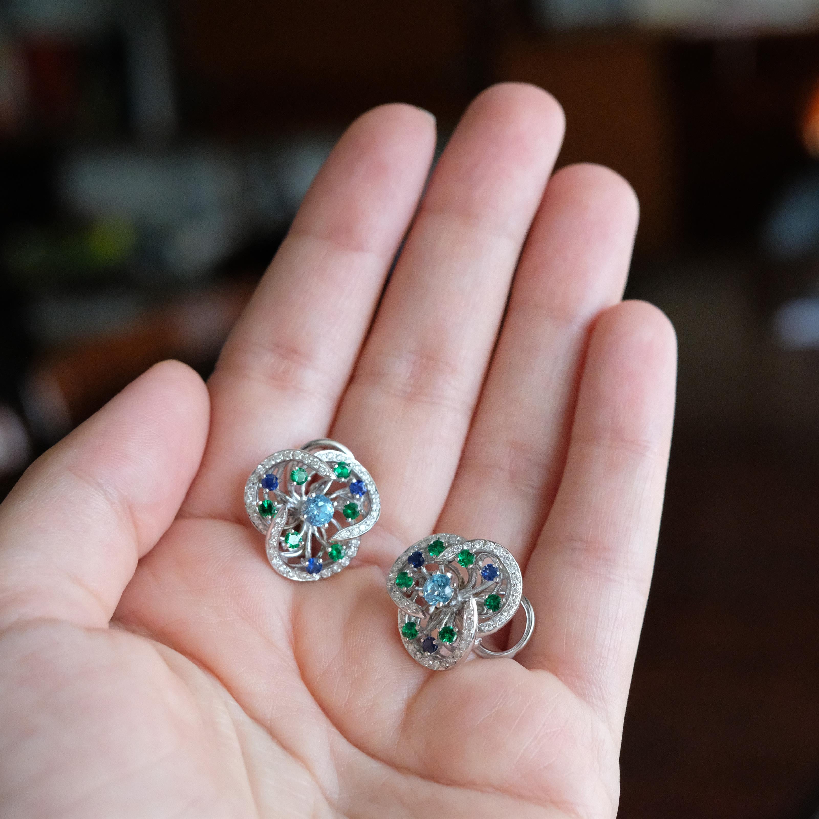 Crafted in white 19.2K gold and set with round blue topaz, white diamonds, tsavorites and blue sapphires, these pretty floral clip-on earrings have the perfect size and a very romantic look. These pieces are designed by Monseo and manufactured by