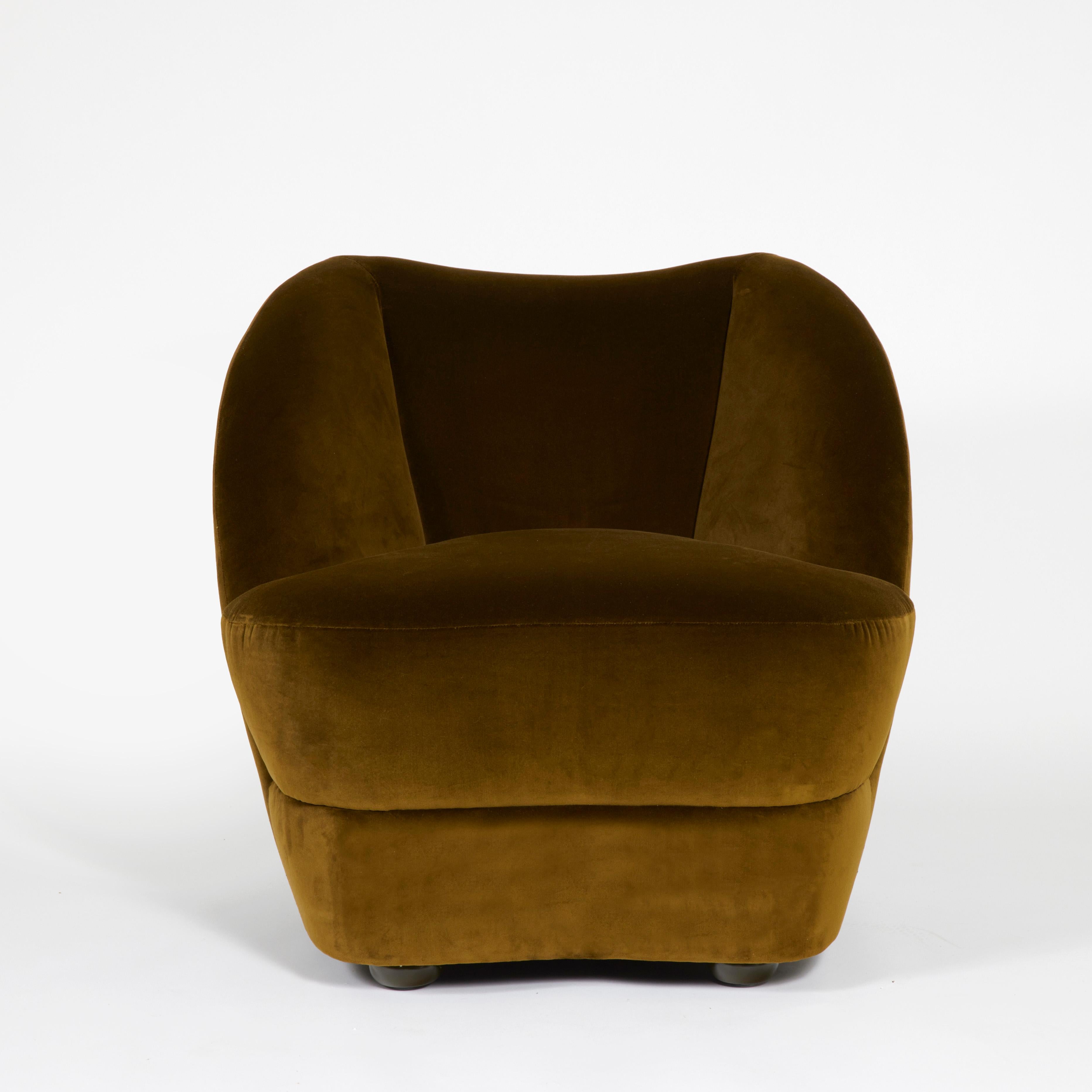 Le Montis est un fauteuil enveloppant au dossier en forme de vague, tapissé de velours et reposant sur quatre pieds boules en bois laqué. Fabriqué avec une structure en chêne et garni de mousse haute densité pour la résilience et de plumes pour un