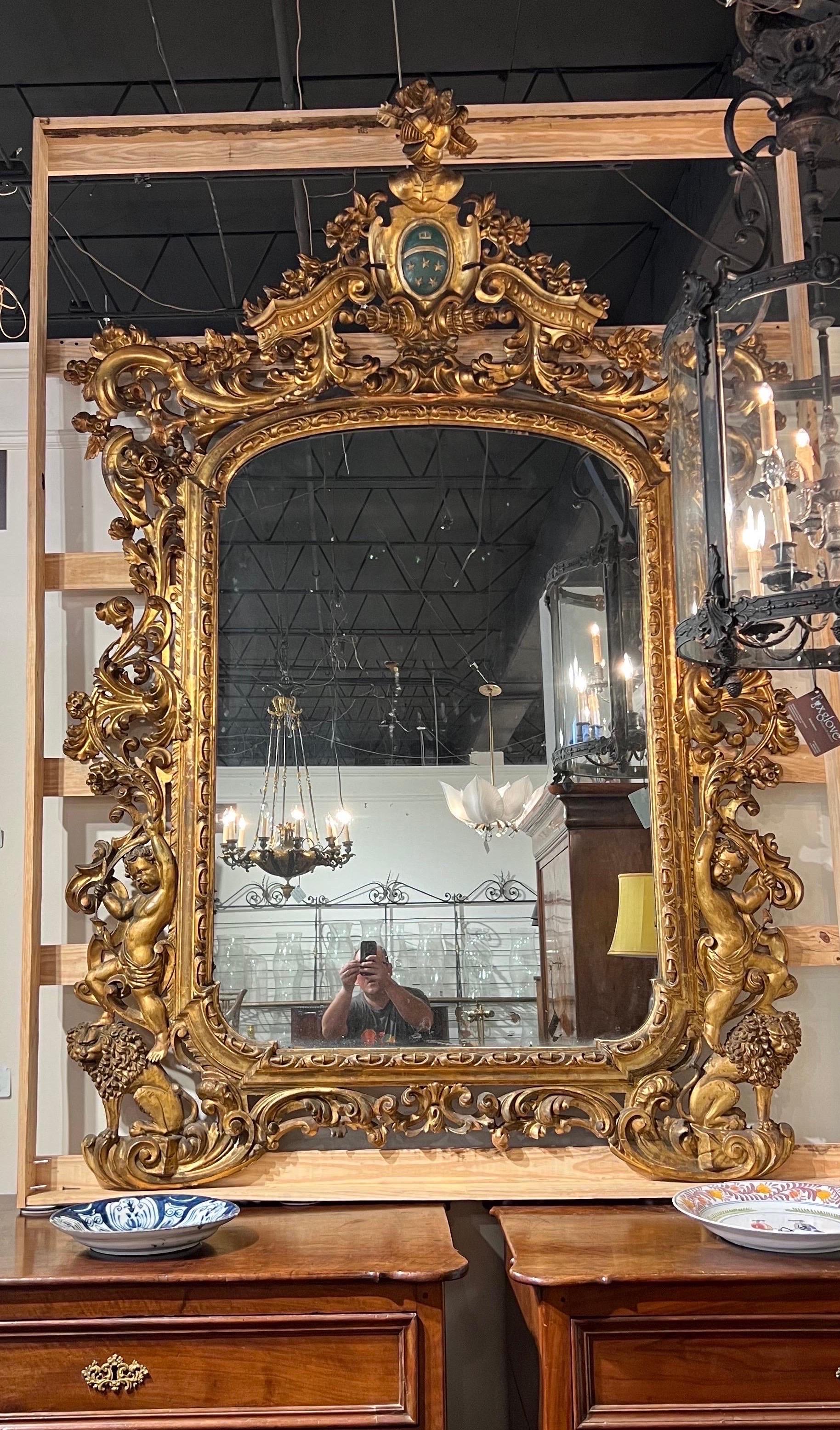 Monumental and impressively over the top 18th-19th century Giltwood mirror, likely Italian. Standing 8’ tall by over 5’ wide, this mirror features heavily carved Giltwood foliage, cherubs, lions and a coat of arms at the crest. Very impressive in