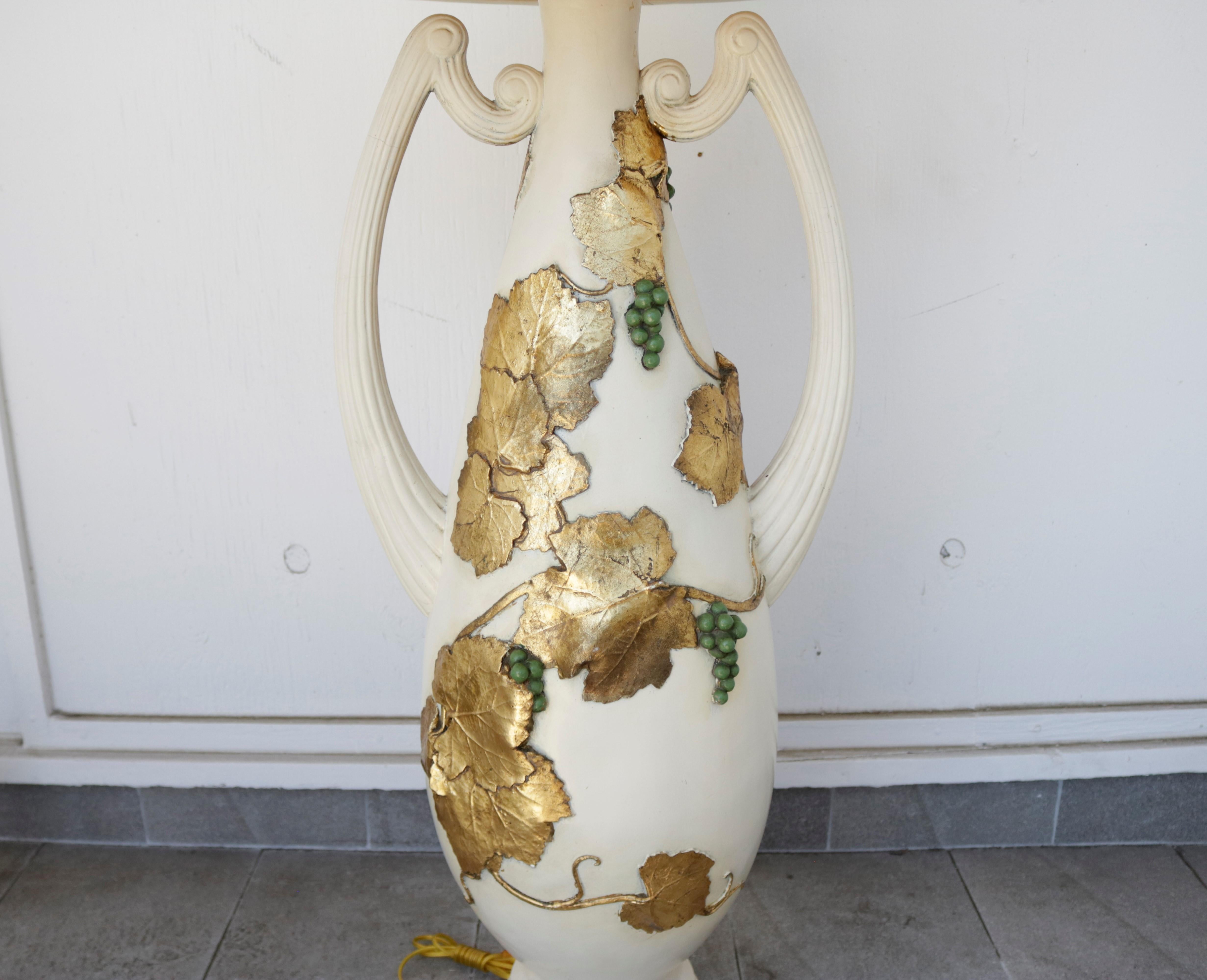 This monumental table lamp features an amphora-shaped base decorated with green grape clusters and twining gold-leafed vines. With an aged ivory glaze finish, the ceramic base measures over 5 feet tall, making this lamp suitable as either a tabletop