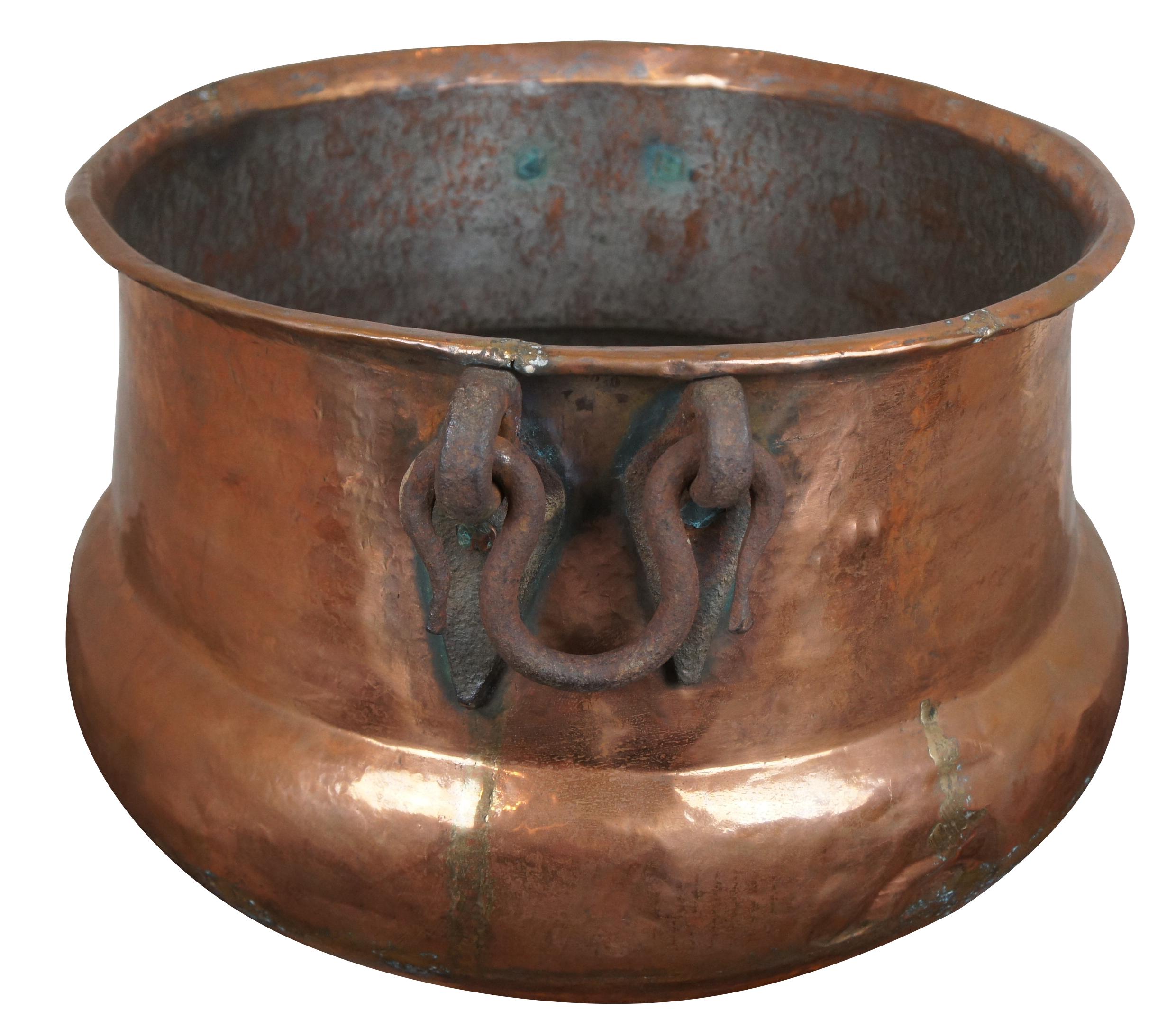 Monumental antique copper cauldron featuring hammered details with dovetailing and ornate forged iron handles.
 