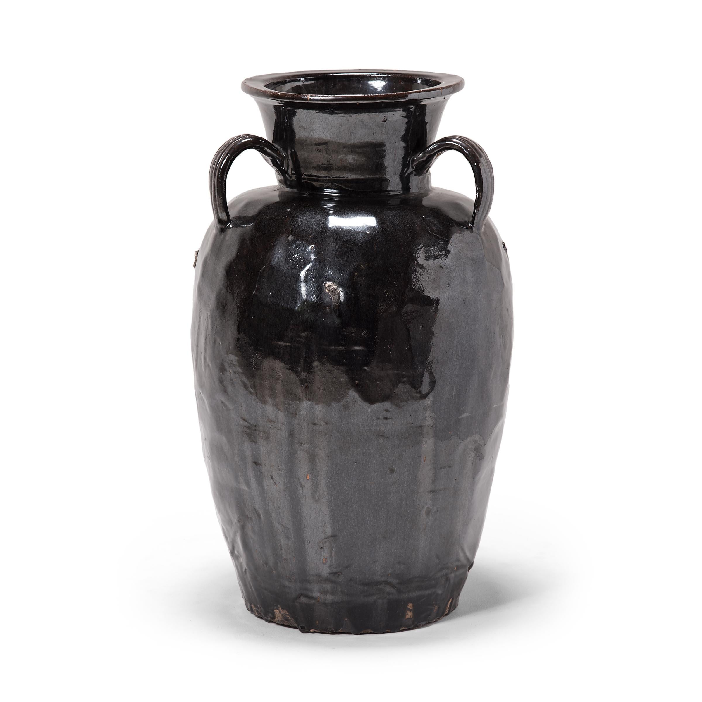 This monumental stoneware vessel was hand-formed in the early 20th century by an artisan in China's Shanxi province. The jar has a wide mouth with a rolled lip and three strap handles attached at the shoulders. A dark, glossy glaze sheets, pools,
