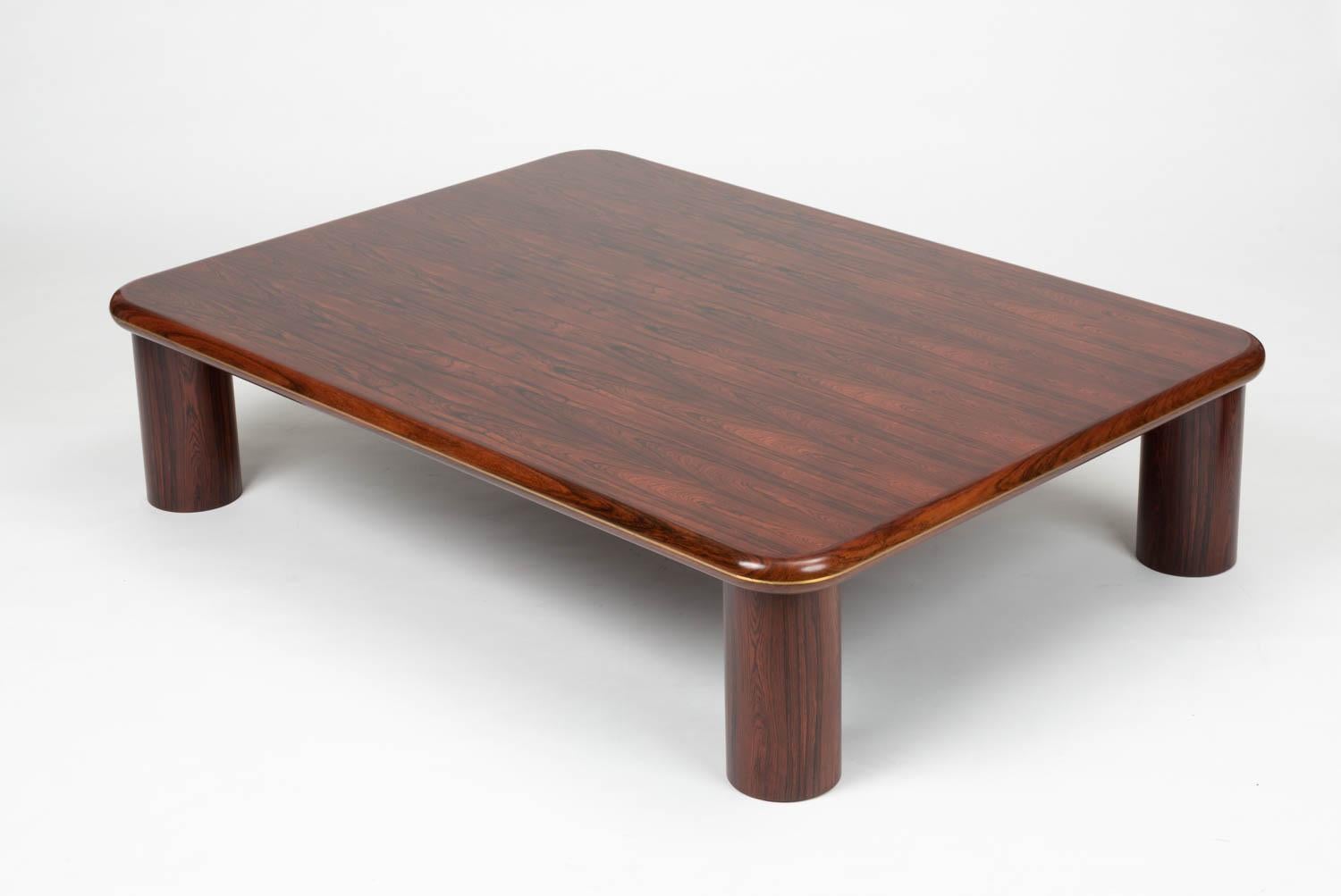 A large-scale coffee table in highly figurative rosewood veneer over hardwood. The solid rosewood banding is accented by a thin brass detail. The thick rectangular top sits atop four round pillar legs, slightly inset from the corners.

Condition: