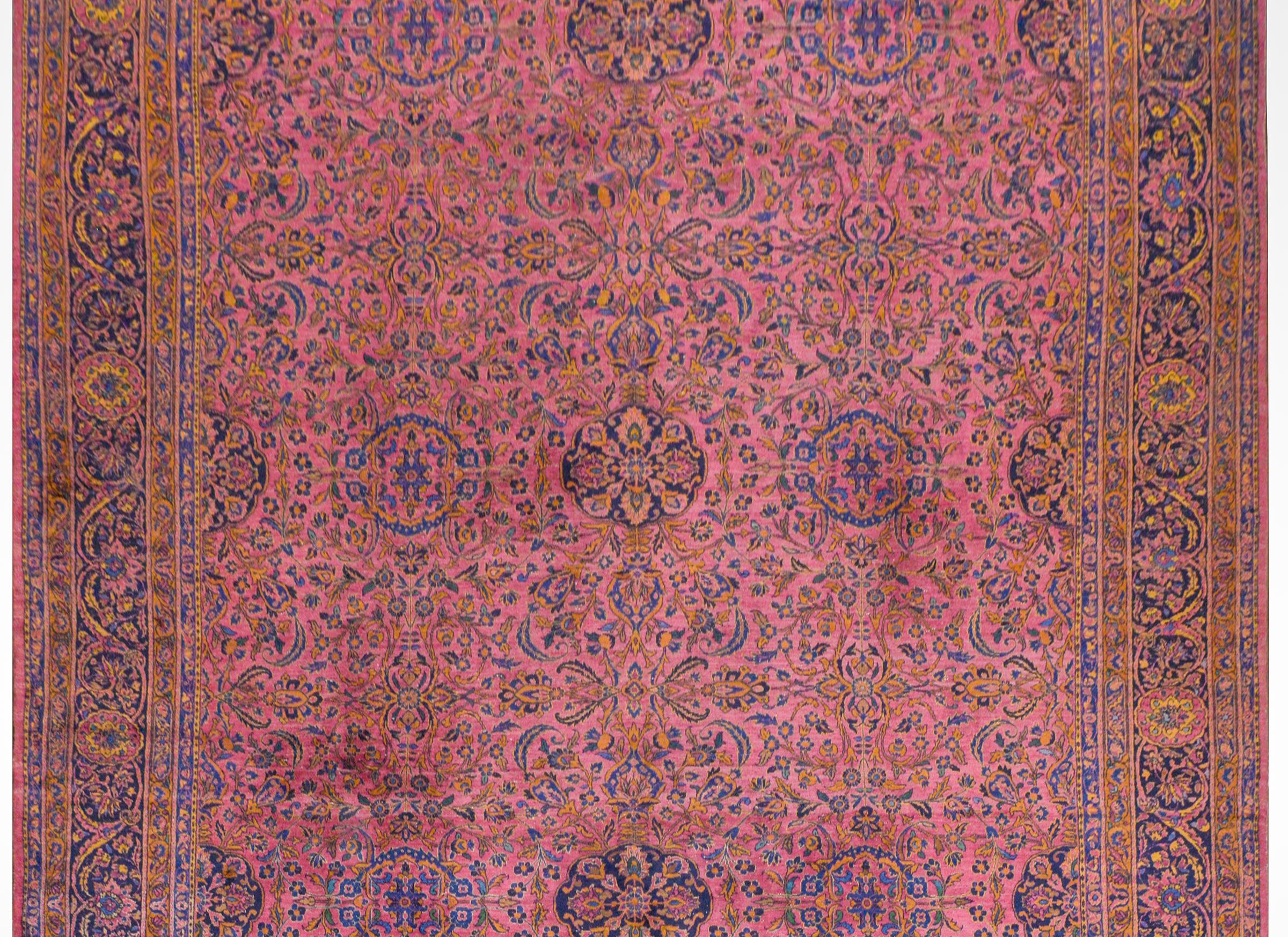 A monumental early 20th century Indian Kashan rug with an all-over floral and vine pattern with multiple floral medallions, all woven in light and dark indigo, gold, and cream colored wool on a bright cranberry background. The border is