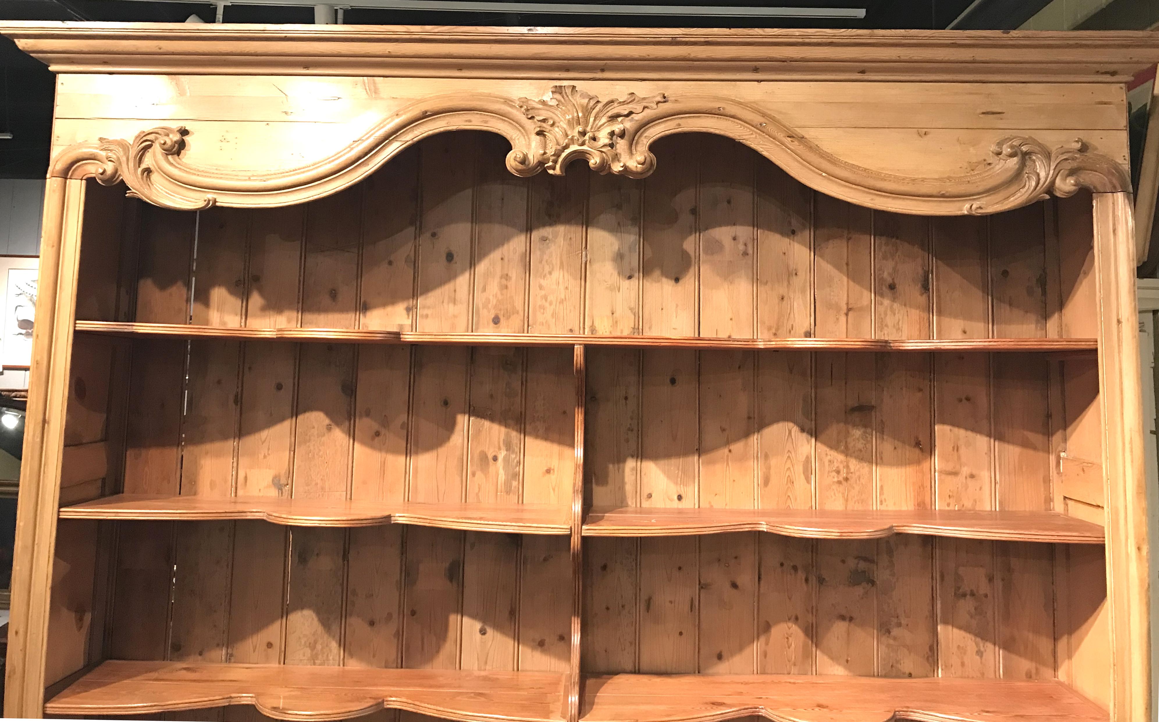 A monumental two part pine bookcase / hutch with foliate carved crest, C and S scroll and acanthus carved decoration, and paneled sides. Its upper case has open shaped shelves with a central divider, and a lower case with three paneled doors with