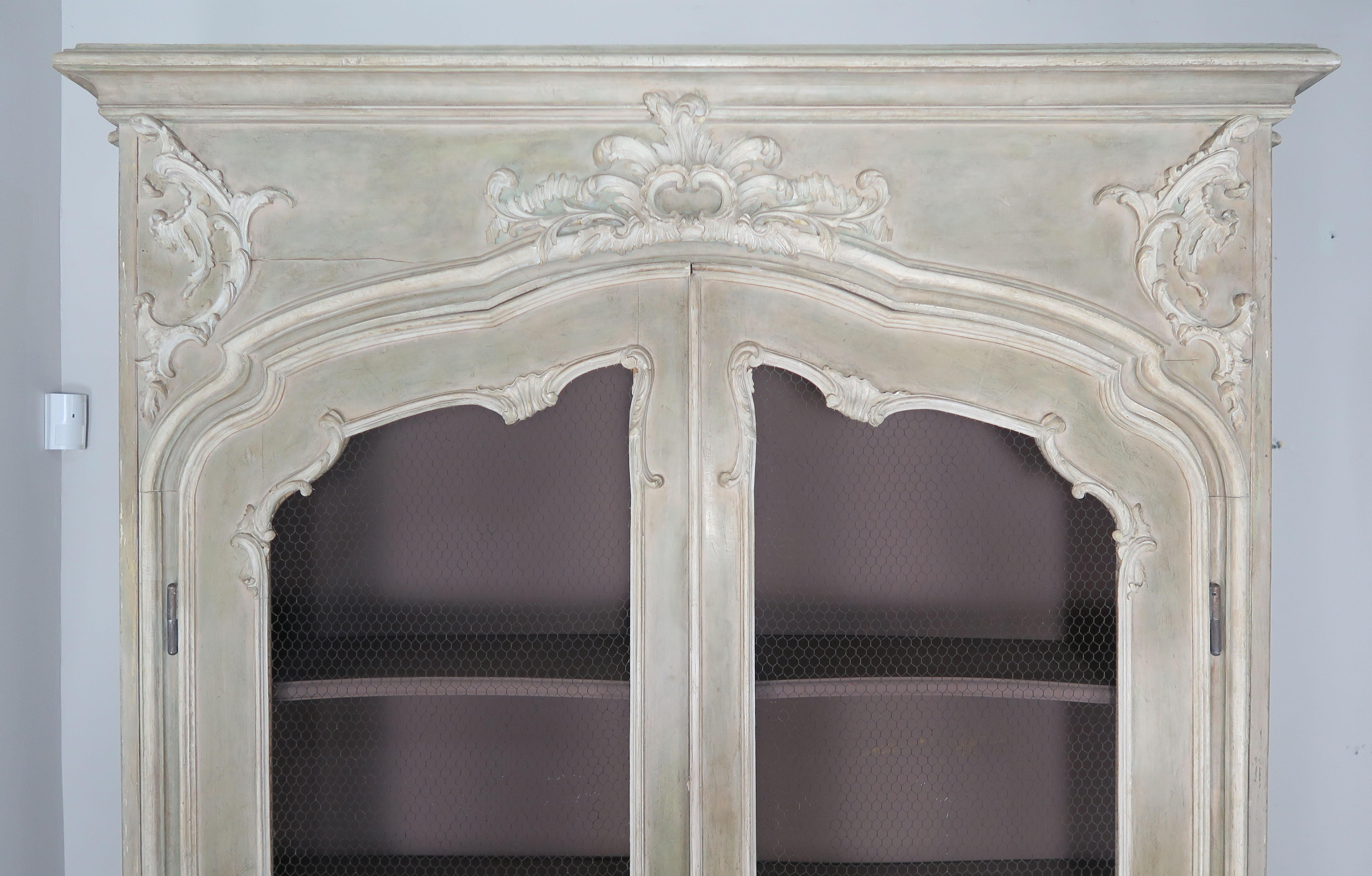 Monumental sized French Louis XV style soft celadon green and cream painted cabinet with scrolls and acanthus leaf detailing. Inset chicken wire doors, shelving and interior lights.