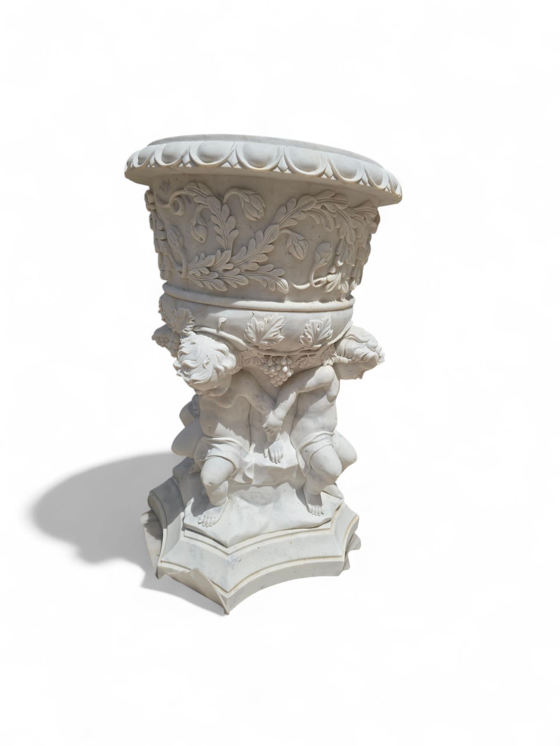 Impressive and elegant monumental urn carved by hand in fine white Carrara marble, featuring charming cherubs (putti) supporting the body of the vessel. The sculptural details, including the figures and decorative motifs, are finely executed,