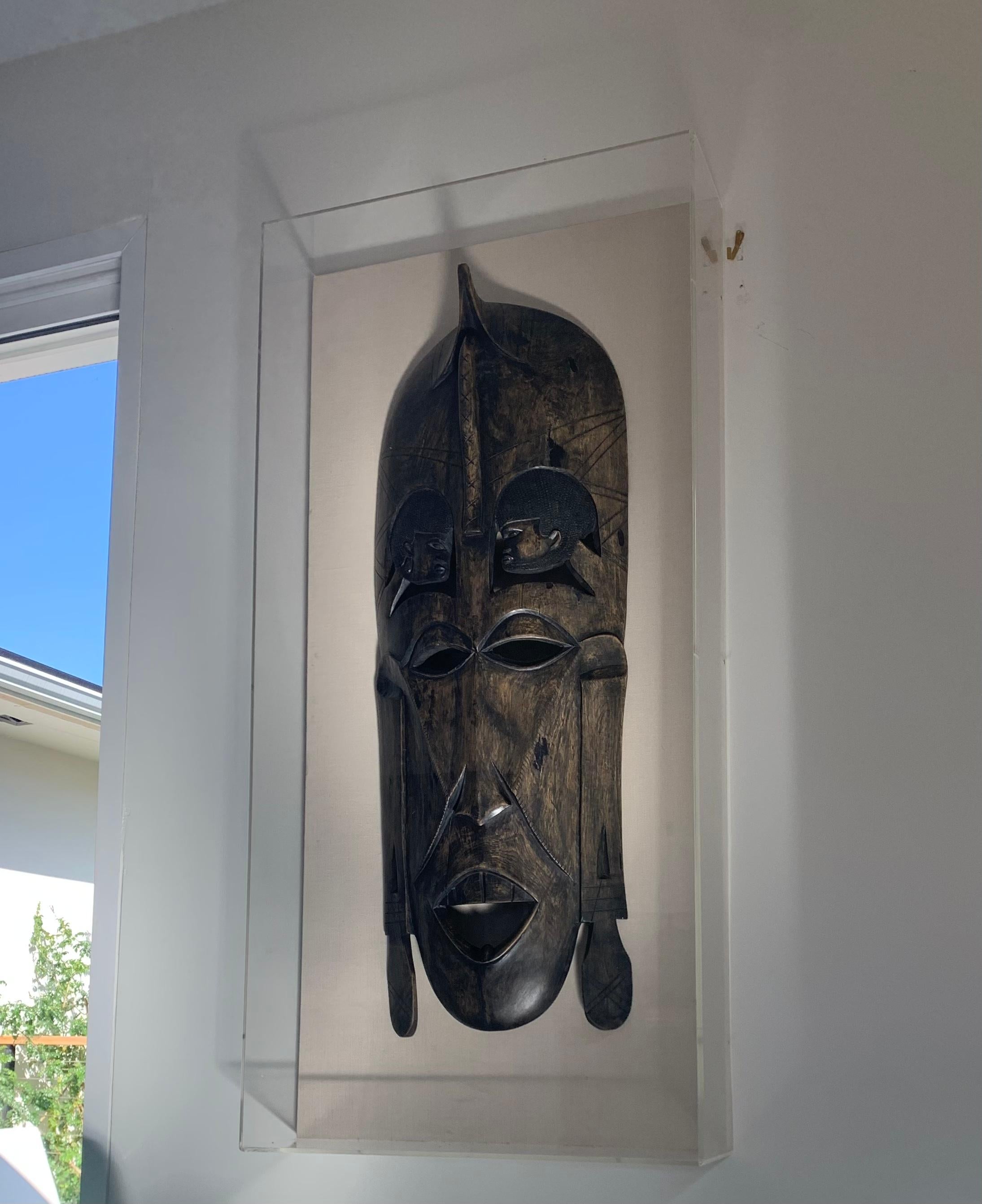 A monumental vintage primitive African mask hand-carved out of mango wood, circa 1960s or possibly earlier. Most likely from Kenya. The mask has been mounted on a linen upholstered wood-stretched canvas, and framed in a lucite box. The frame is also