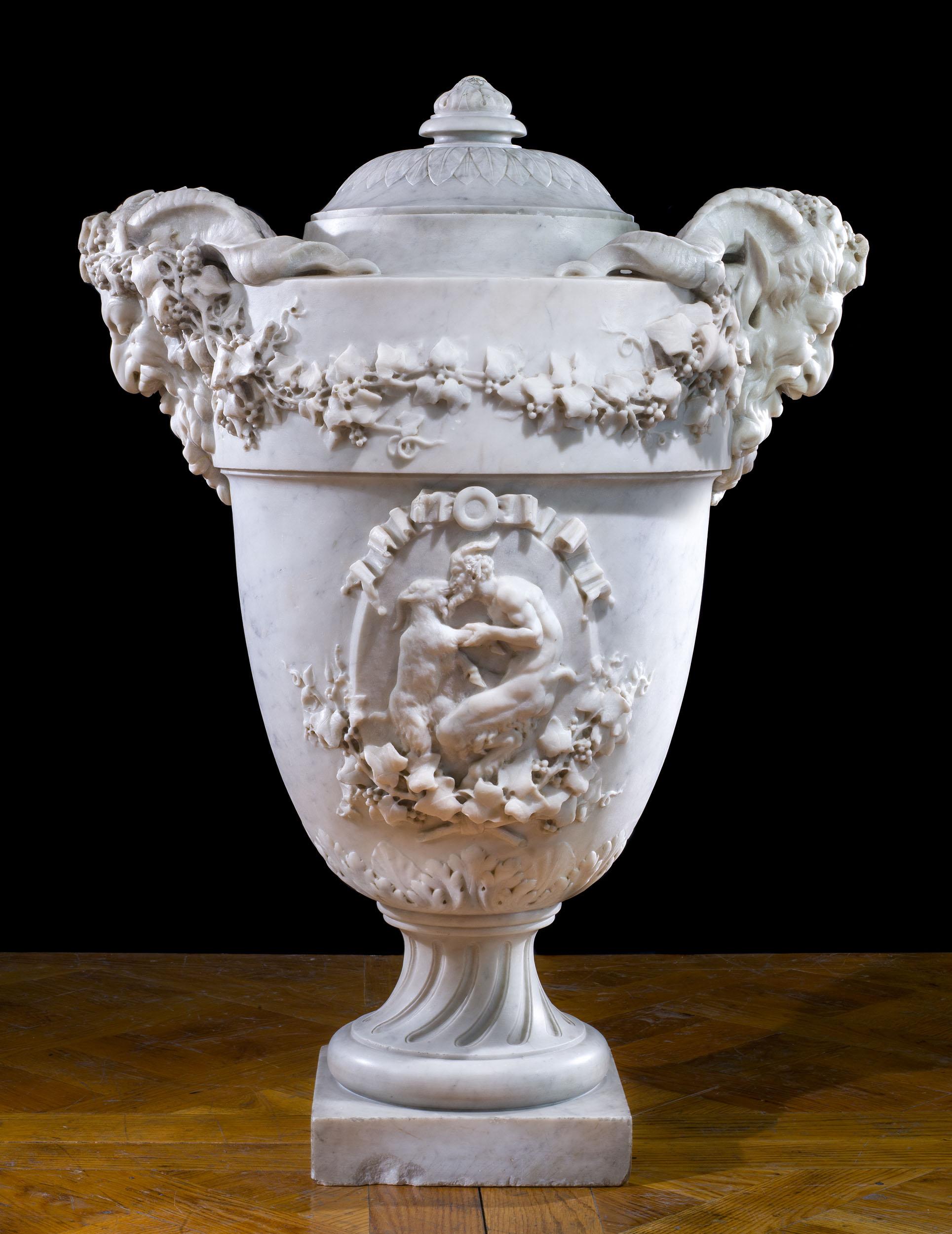 A large and finely carved 19th century Italian marble lidded urn, decorated with trailing grape vines and and dramatic handles, modelled as grinning and horned satyrs. The body of the urn is carved with two cartouches, one depicting a nymph bathing