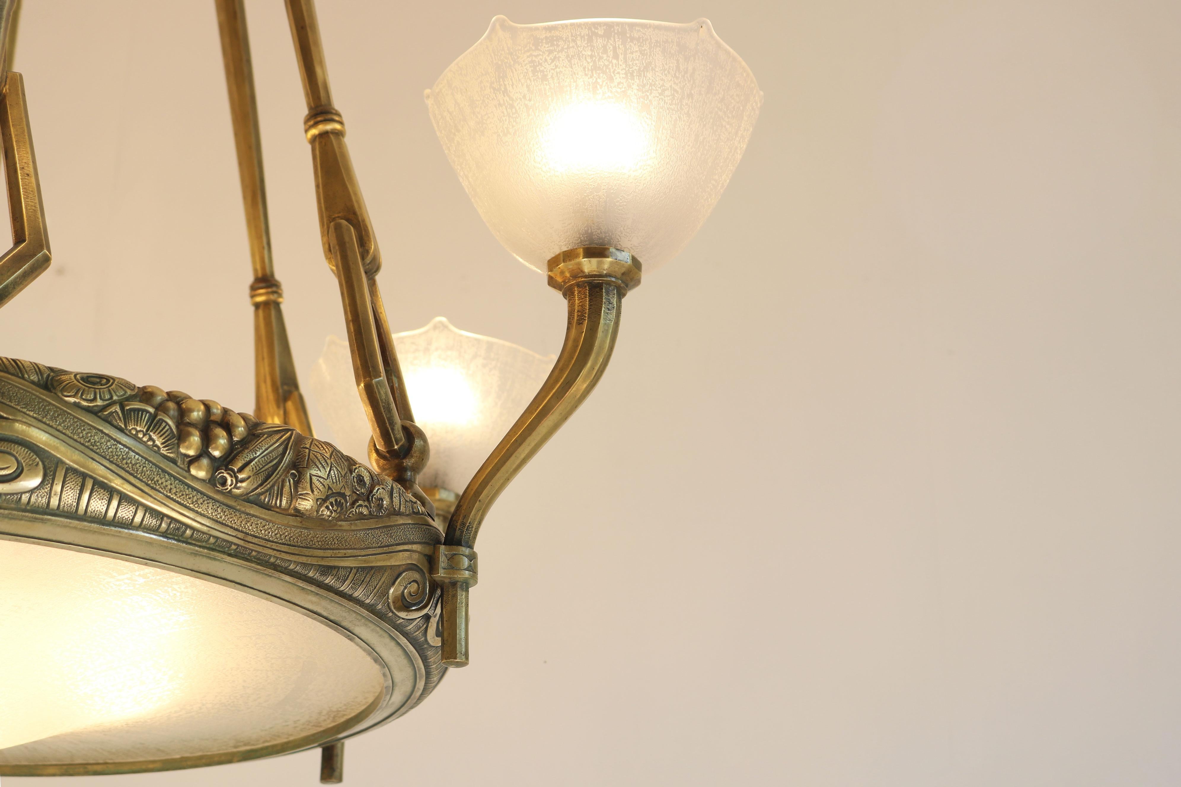 Exquisita araña antigua Art Déco, una obra maestra monumental diseñada por Maurice Dufrene en 1925. Presenta seis elegantes brazos con doce luces luminosas, expertamente elaborados en bronce dorado. El marco está adornado con llamativos motivos