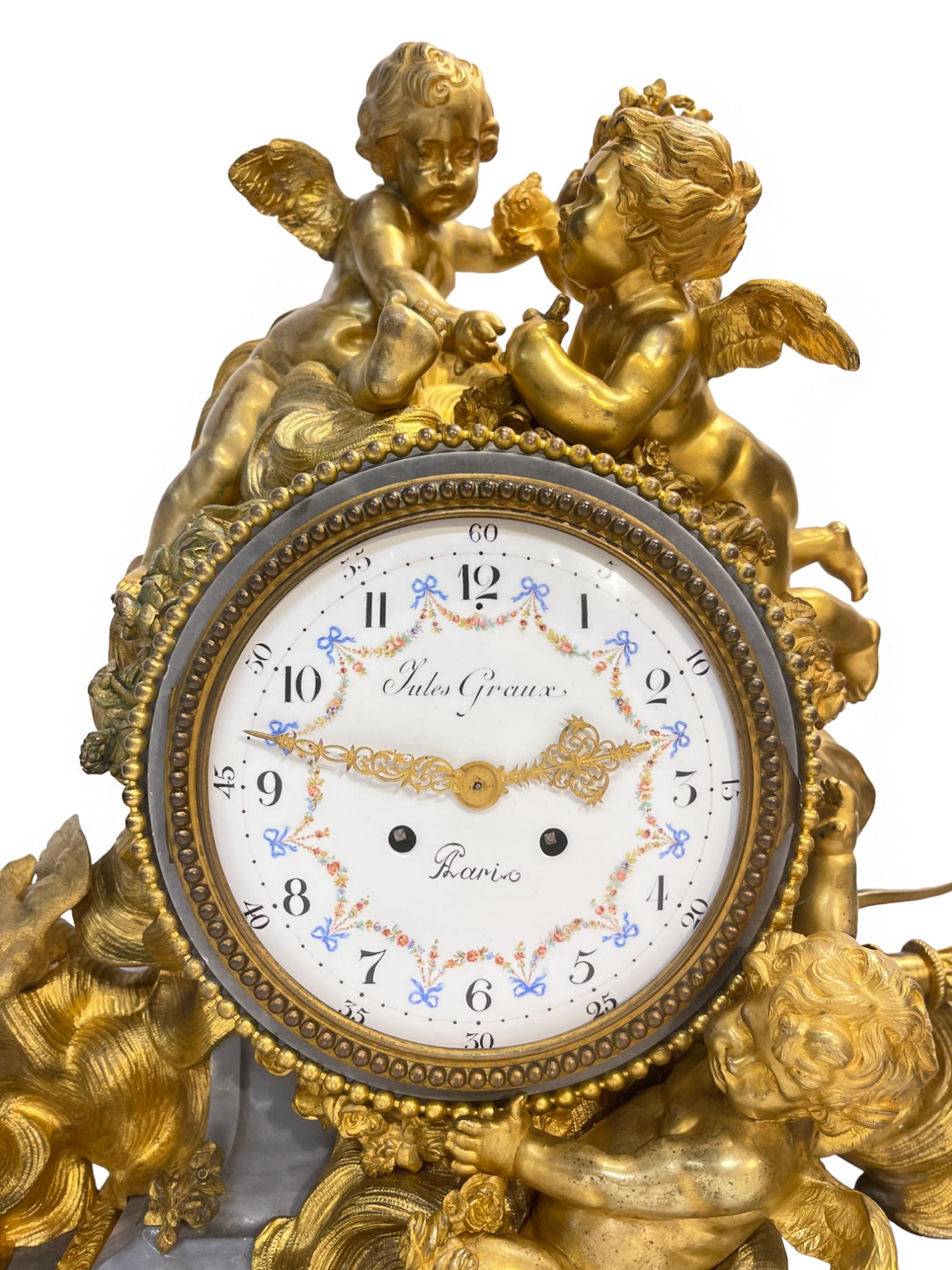 A fine Louis XVI-style large gilt-bronze and greige Rococo marble figural mantel clock is surmounted by three dore bronze putti figures hanging floral adornments among clouds with love birds and Cupid's bow. Raised on bronze feet with ormolu