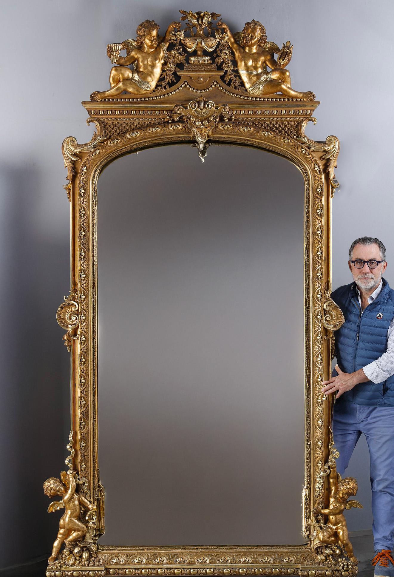 Majestic mirror featuring a richly carved and gilded frame decorated with scrolls, pearl friezes, acanthus leaves and shells. The pediment is adorned with two musician cherubs in the round, on either side of a flowery cassolette, each holding a