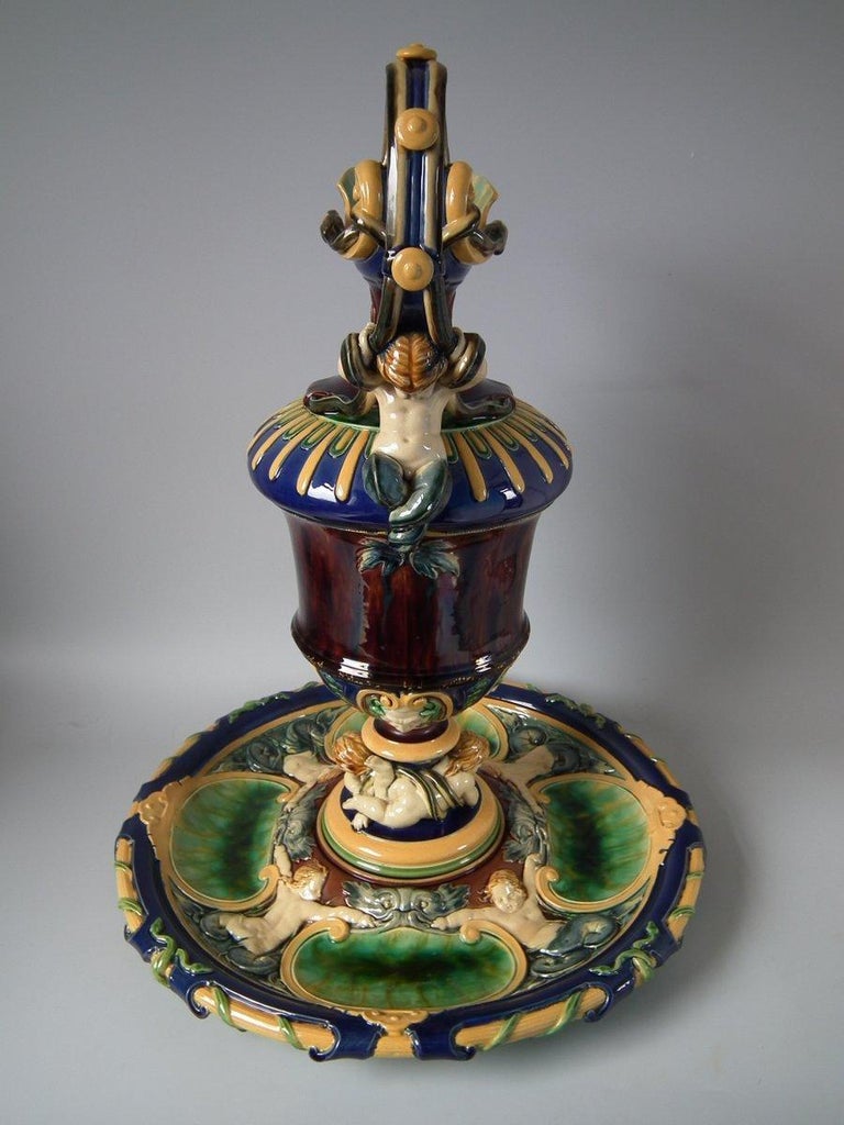 Minton Majolica ewer and stand which features putti supporting the stem of the ewer, a cherub riding a dolphin below the spout. A merboy with entwined serpents to the handle. The base has serpents around the rim. Merboys and satyr heads with open