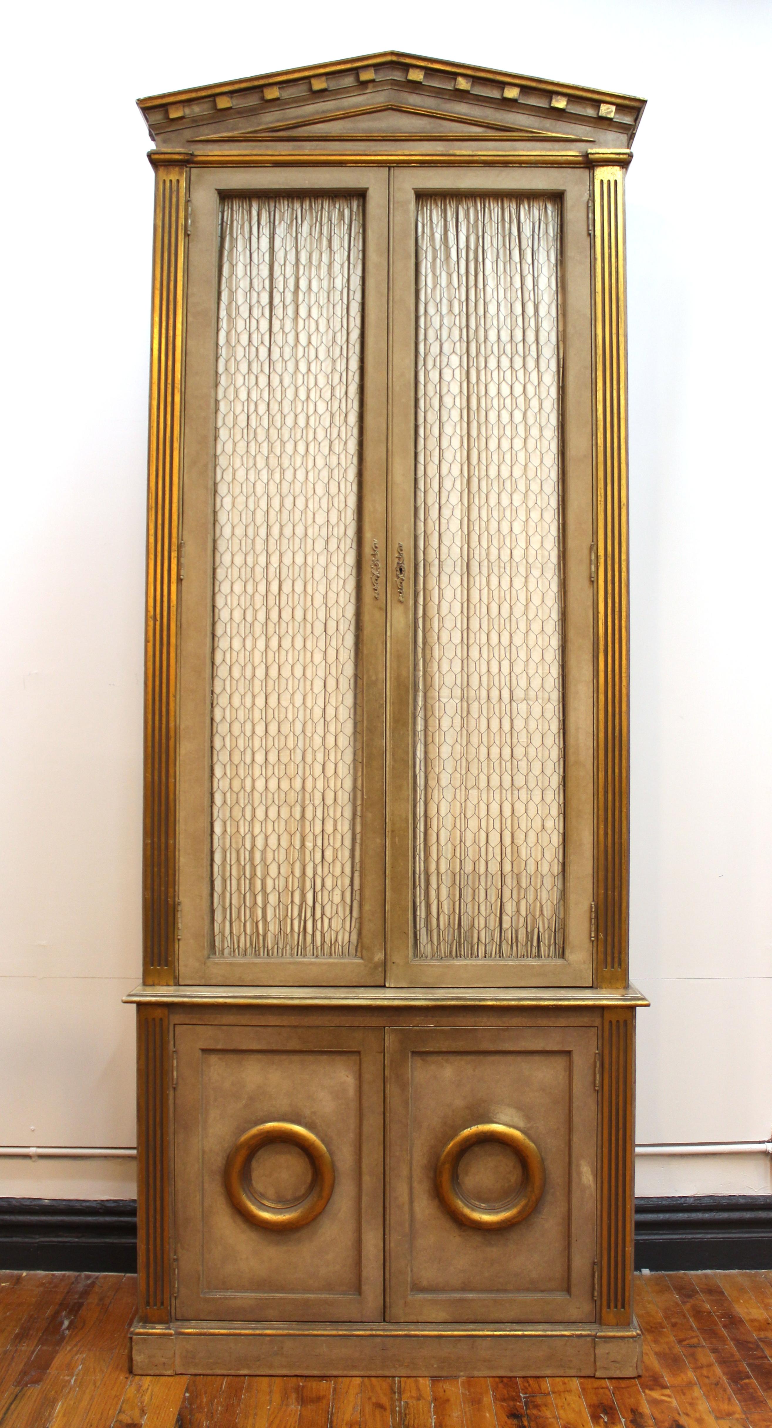 Monumental pair of wood cabinets in Neoclassical Revival style with pedimented tops. The pair has gilt accents and the upper parts can be separated easily from the lower cabinets. The doors of the upper section have wire in the opening and textile