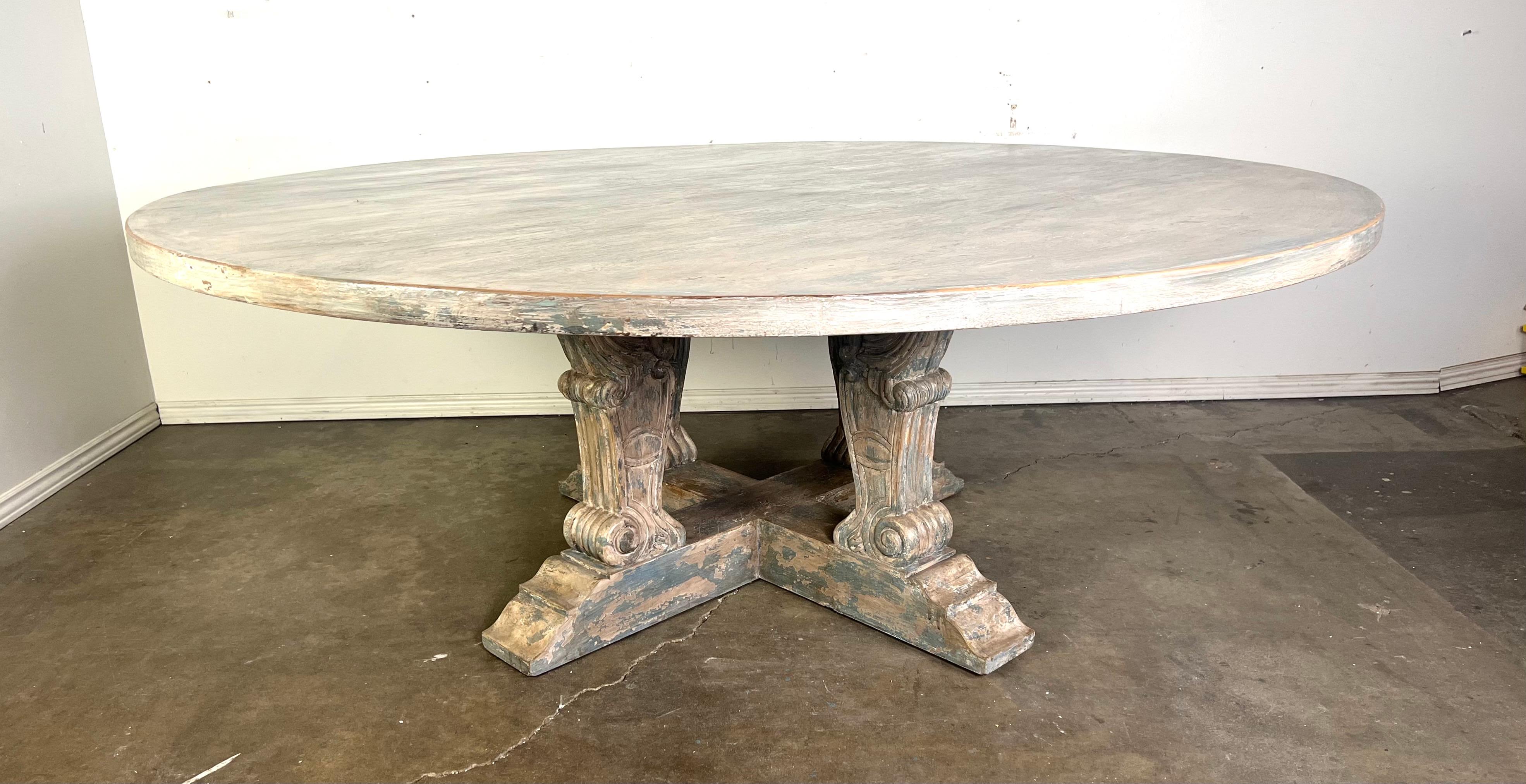 Table de salle à manger ronde de style néoclassique, de taille monumentale, à la finition rustique et vieillie.  Son design se caractérise par des pieds piédestaux sculptés de manière complexe, avec des motifs décoratifs de volutes et de feuilles