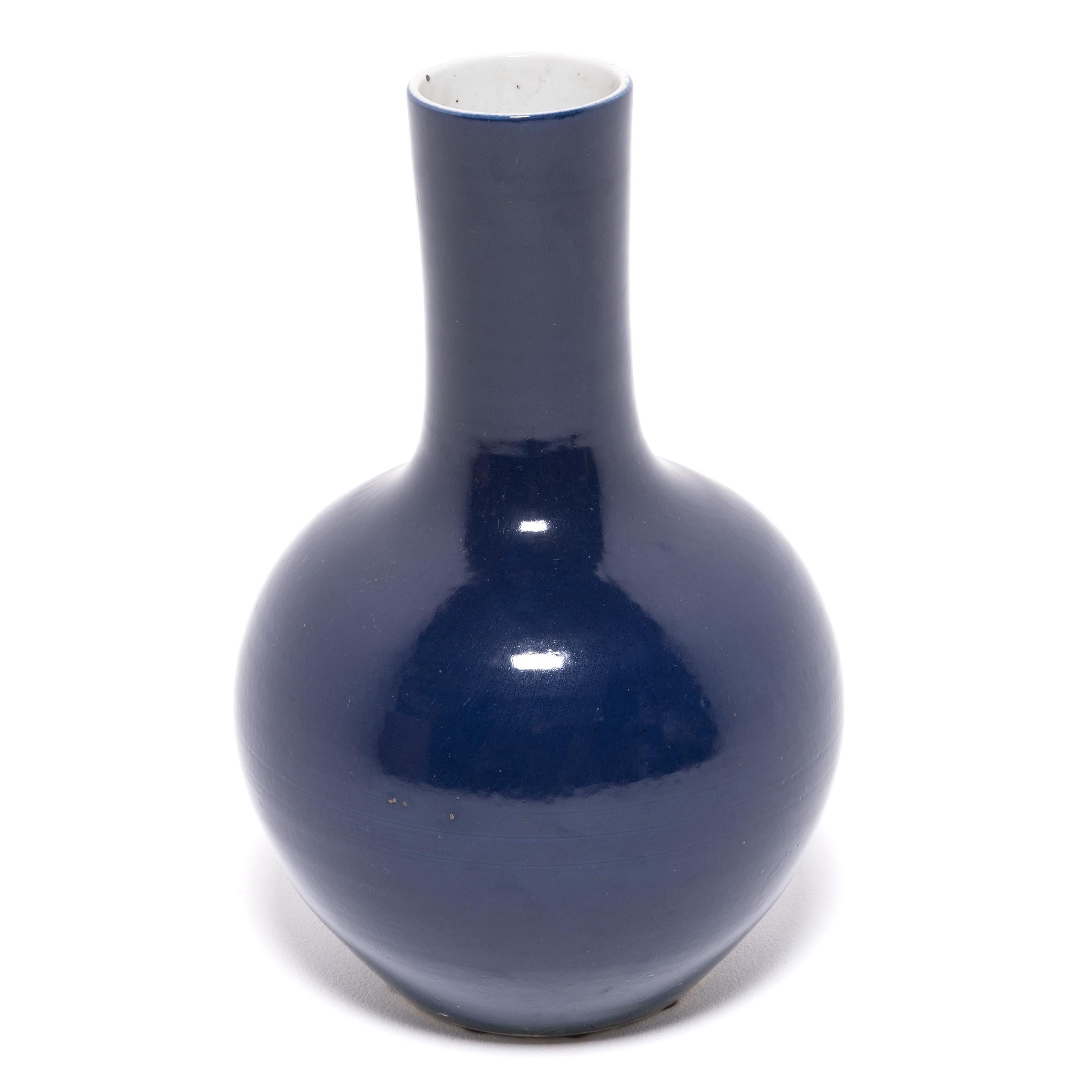 Drawing on a long Chinese tradition of monochrome ceramics, this tall gooseneck vase is coated in a deep blue glaze, reminiscent of a dark night sky. The vase features a rounded, globular body and a narrow cylindrical neck, a classic form known as