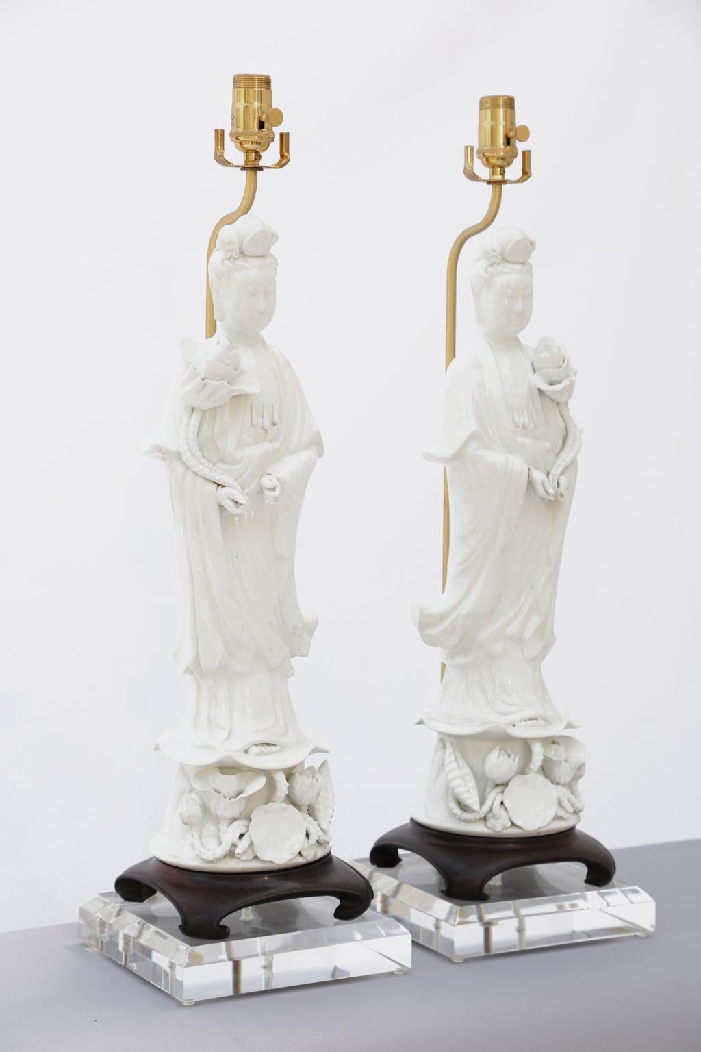 Opposing pair of very large, Blanc de Chine porcelain figures, each Kwan Yin goddess holding a large blossom, standing on a plinth of flowering vines, raised on a wooden base. The detachable figures are lamped on acrylic platforms, fitted for