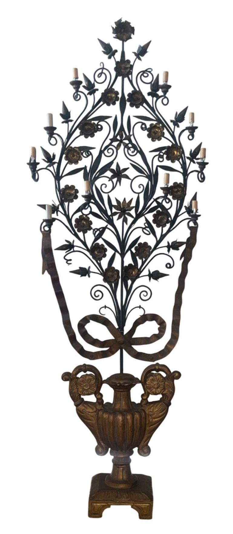 A dramatic pair of tall Italian girandoles, each conceived as a stylized flowering tree rising from an urn. The wrought‑iron frames are enriched with gilt tole blossoms, leaves, and ribbon bows, the branches issuing multiple candle‑form lights and