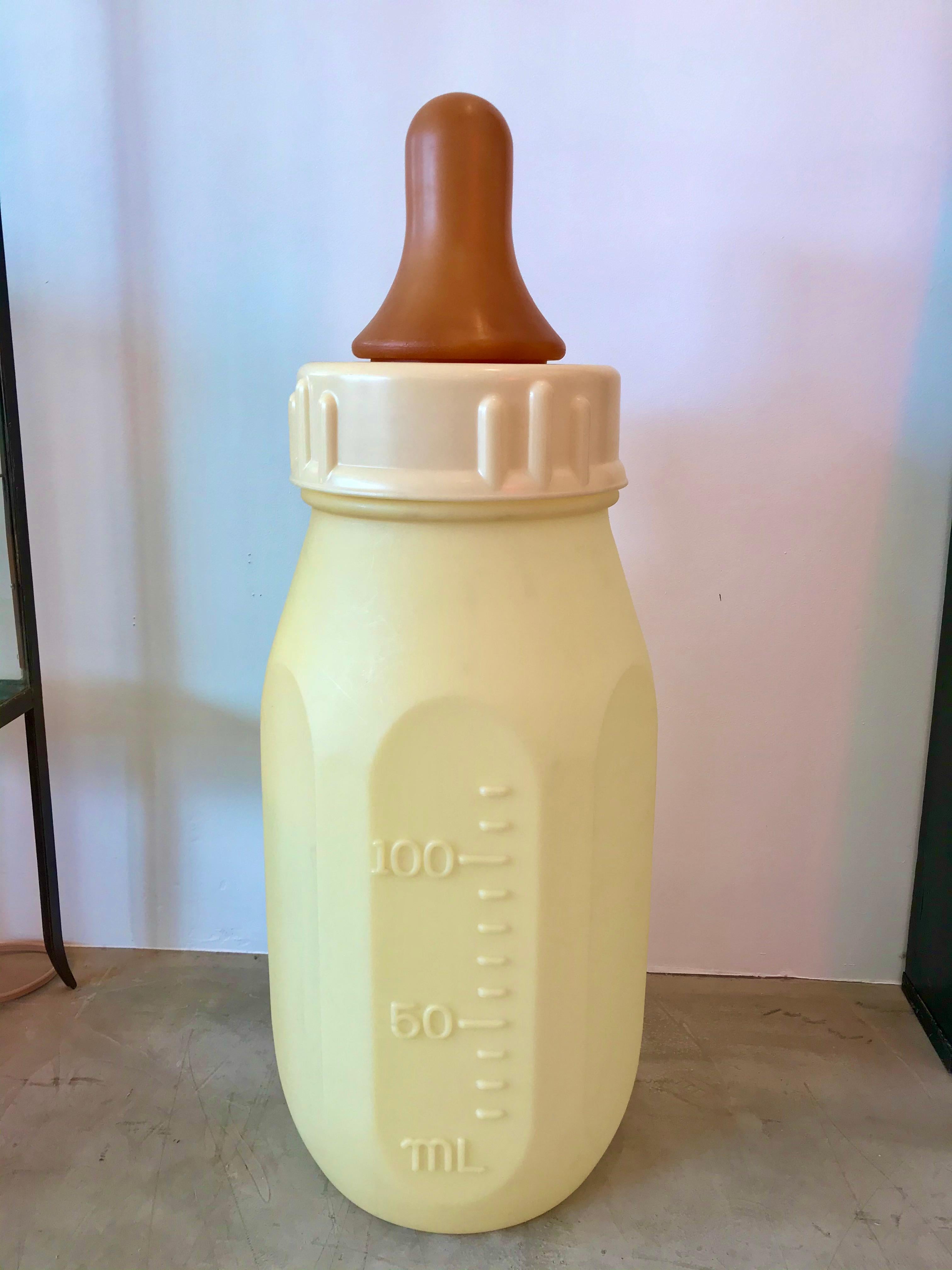 Gigantic baby bottle made in the 1970s. Hard plastic bottle with removable top and rubber nipple. Pale light yellow bottle with ounces on one side and milliliters on the other. At three and a half feet tall and 15