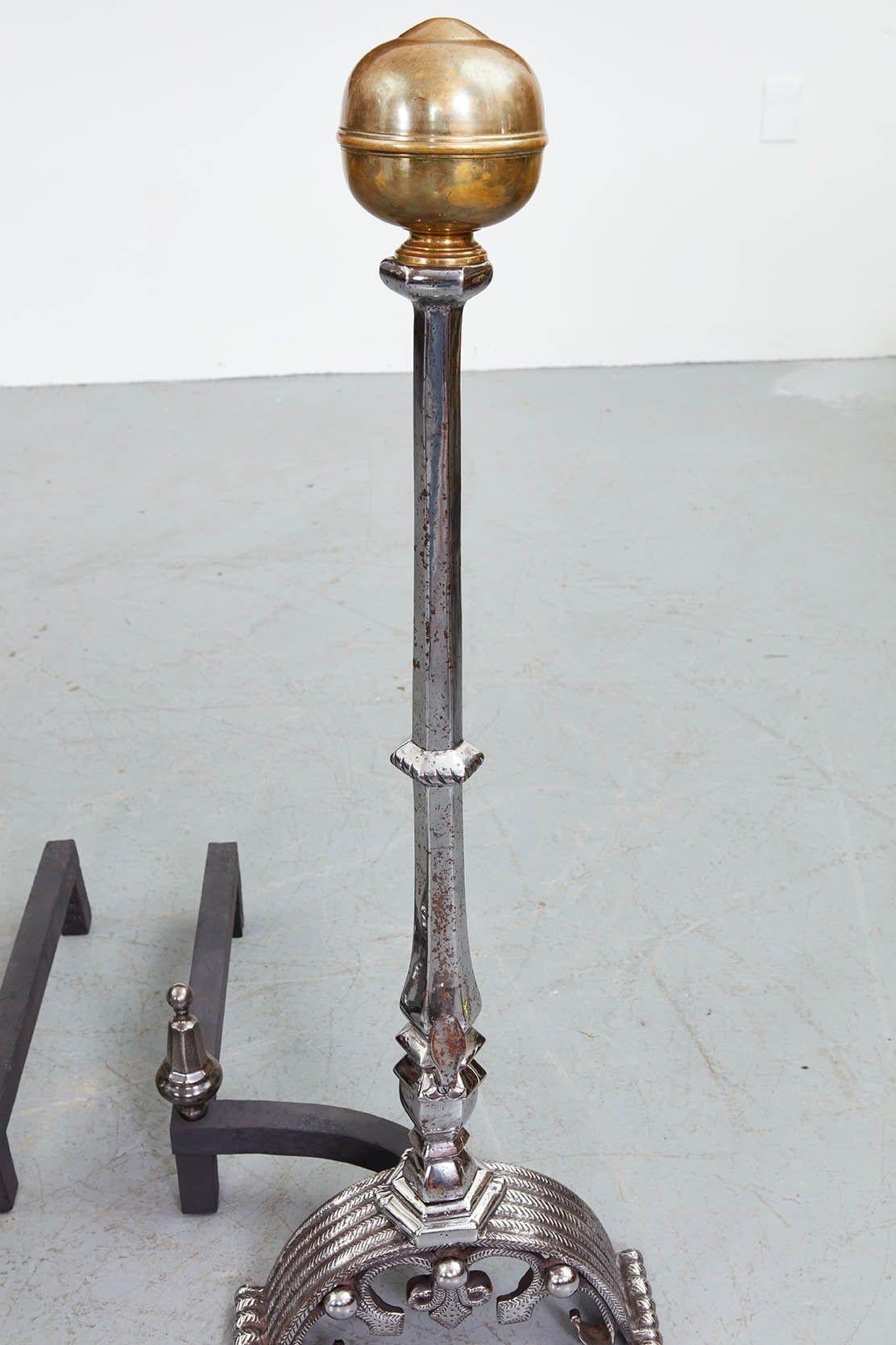 A pair of monumental and impressively scaled andirons with tall faceted polished steel shafts on substantial striated and decorated arched bases, having steel finials on back supports, and surmounted by seamed brass ball finials.