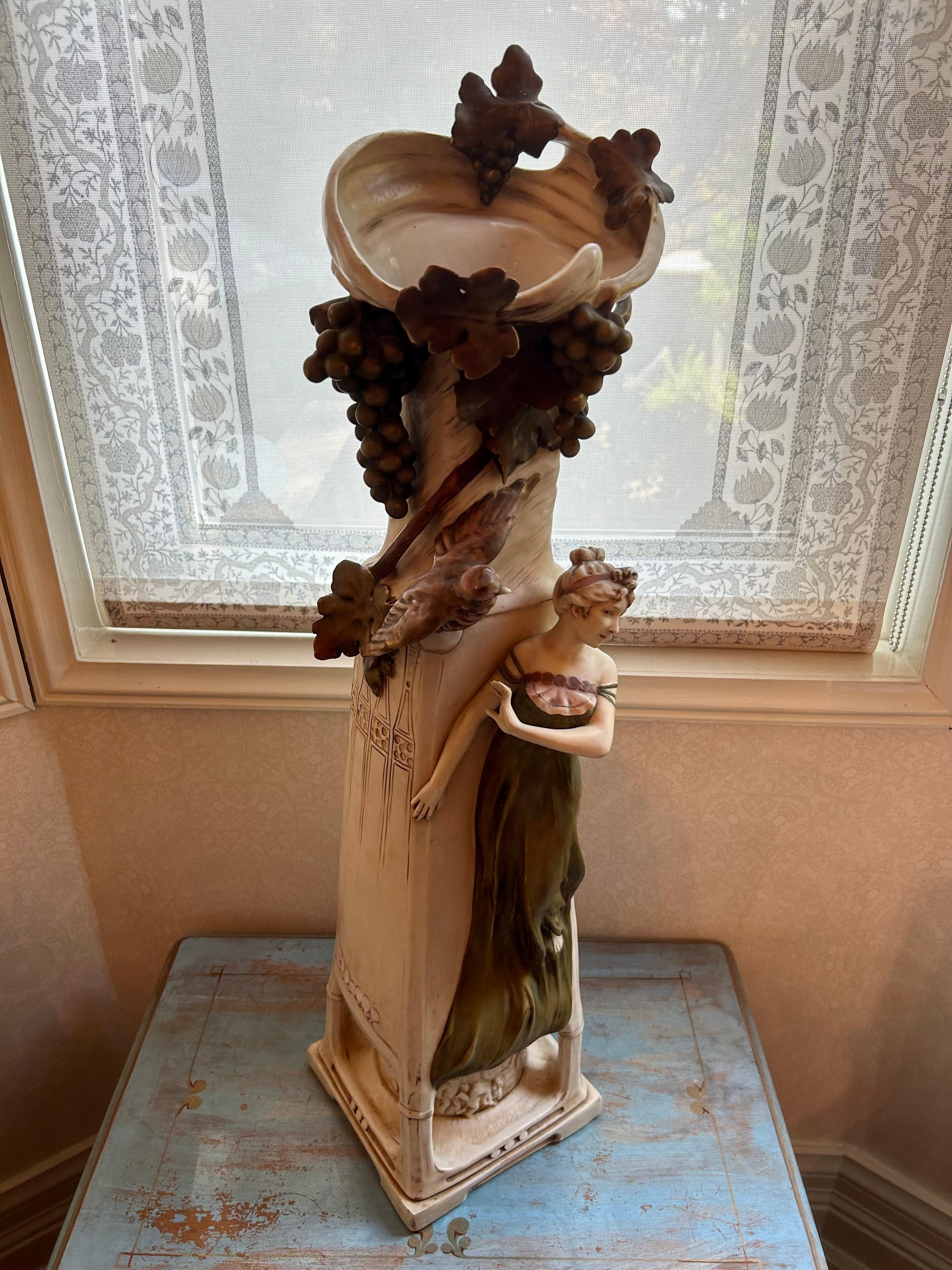 A monumental Royal Dux Bohemia porcelain vase, featuring a beautiful maiden standing under a grapevine, with a large bird. The base of the vase featuring frolicking putti. Typical Art Nouveau motifs throughout, stylized incised lines and pale pink