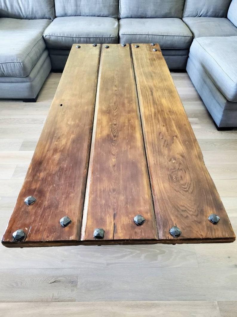 A one-of-a-kind coffee table, handcrafted, the rustic wood reclaimed from an antique door, having heavy industrial hammered stud accents, pine plank wooden top, rich antique wood color and patina, rising on x shape iron legs with single cross