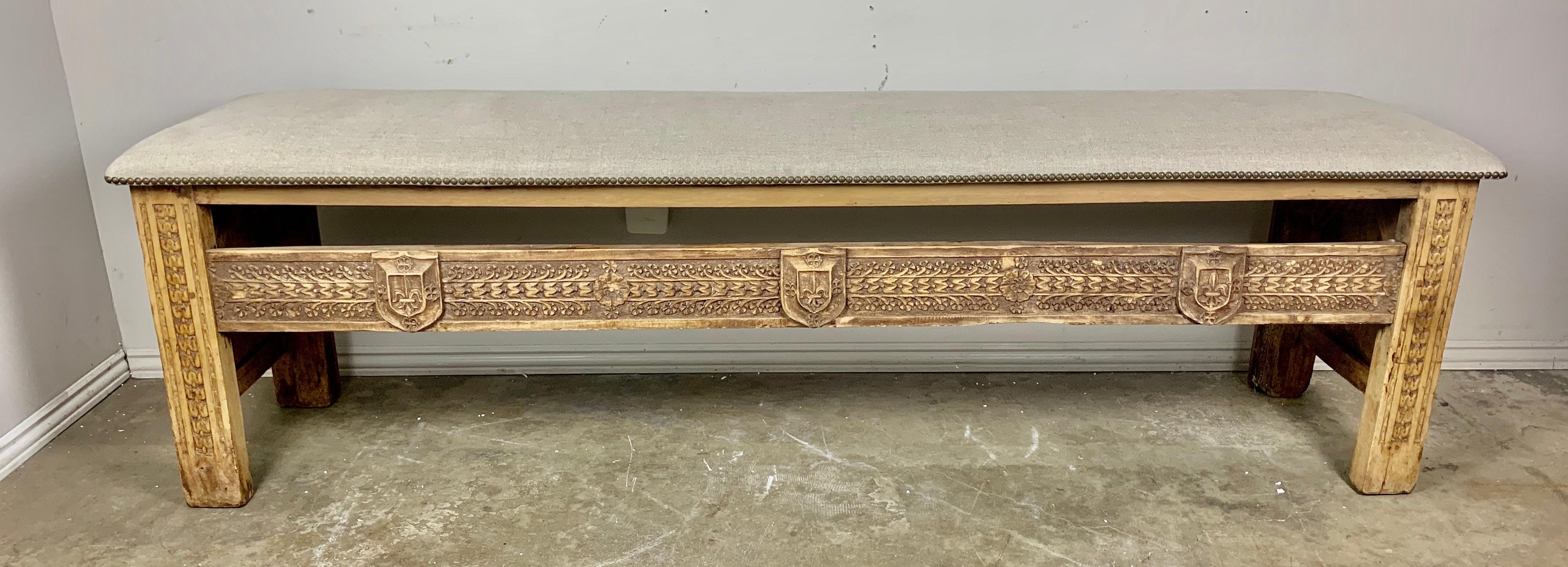 Early 19th century Spanish carved wood bench depicting beautiful detailed shields, fleur dys lis, and so much more. This monumental scaled bench has been newly upholstered in Belgium linen with antique brass nailheads.